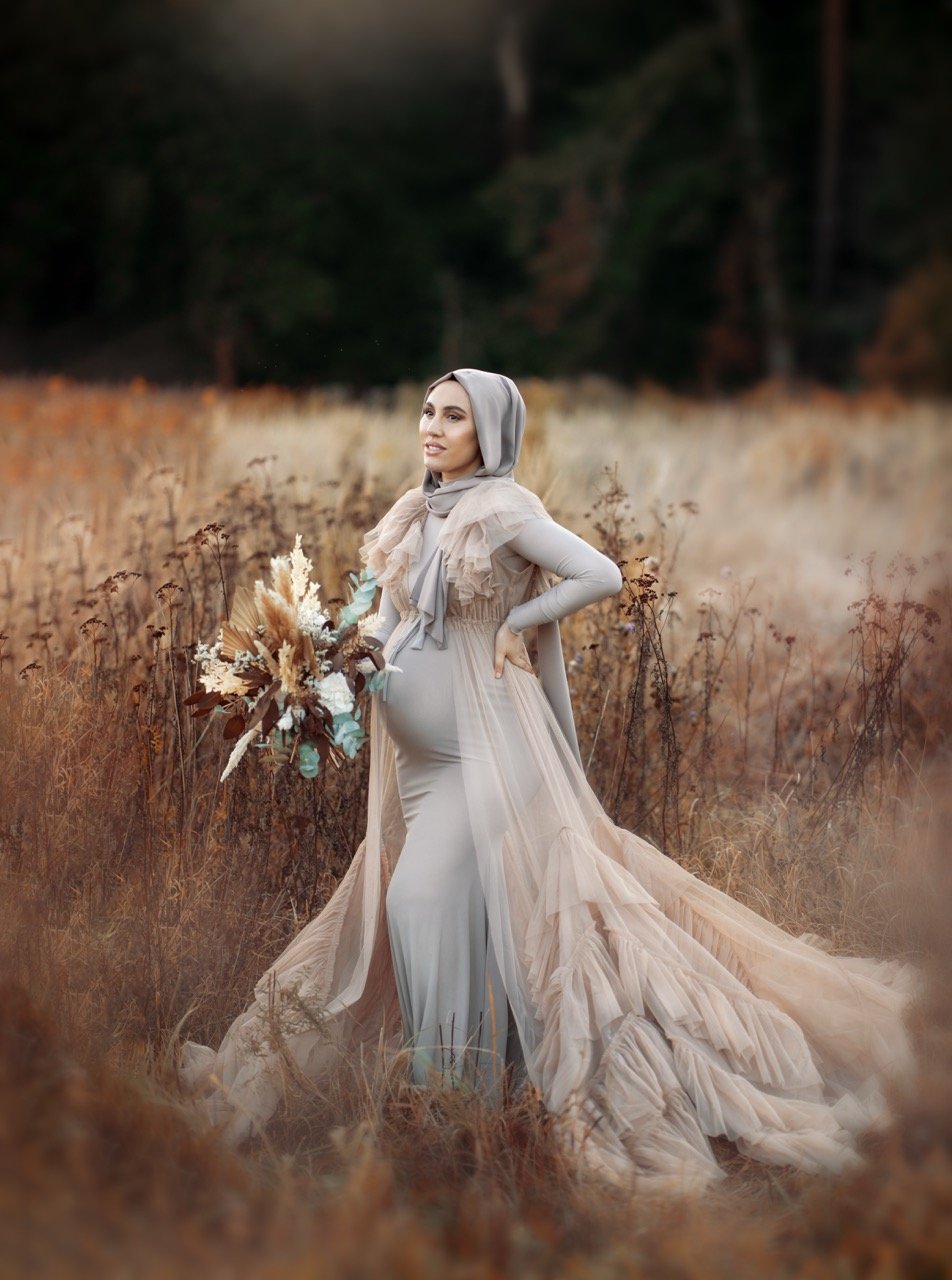 Schwangere Frau im Hijab in einem schicken langen Kleid steht auf einem Feld mit einem Blumenstrauß in der Hand.