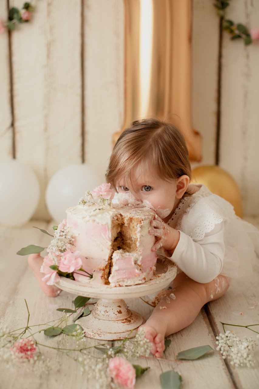 Ein Kleinkind in einem weißen Kleid kniet vor einem rosa dekorierten Kuchen und steckt sein Gesicht hinein. Blumen und Ballons sind im Hintergrund zu sehen.