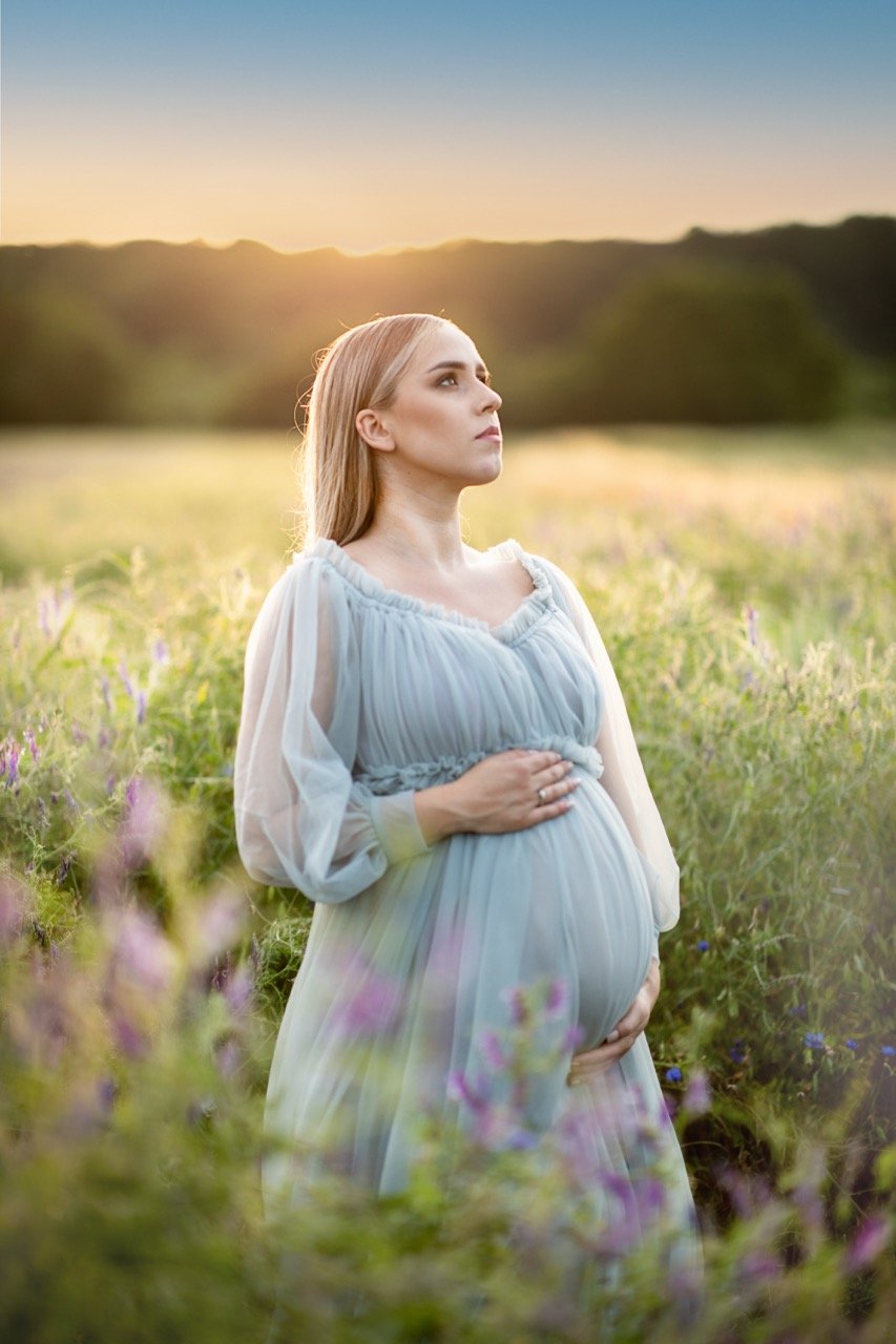 Schwangere Frau in einem blauen Kleid steht auf einem Feld bei Sonnenaufgang oder Sonnenuntergang.