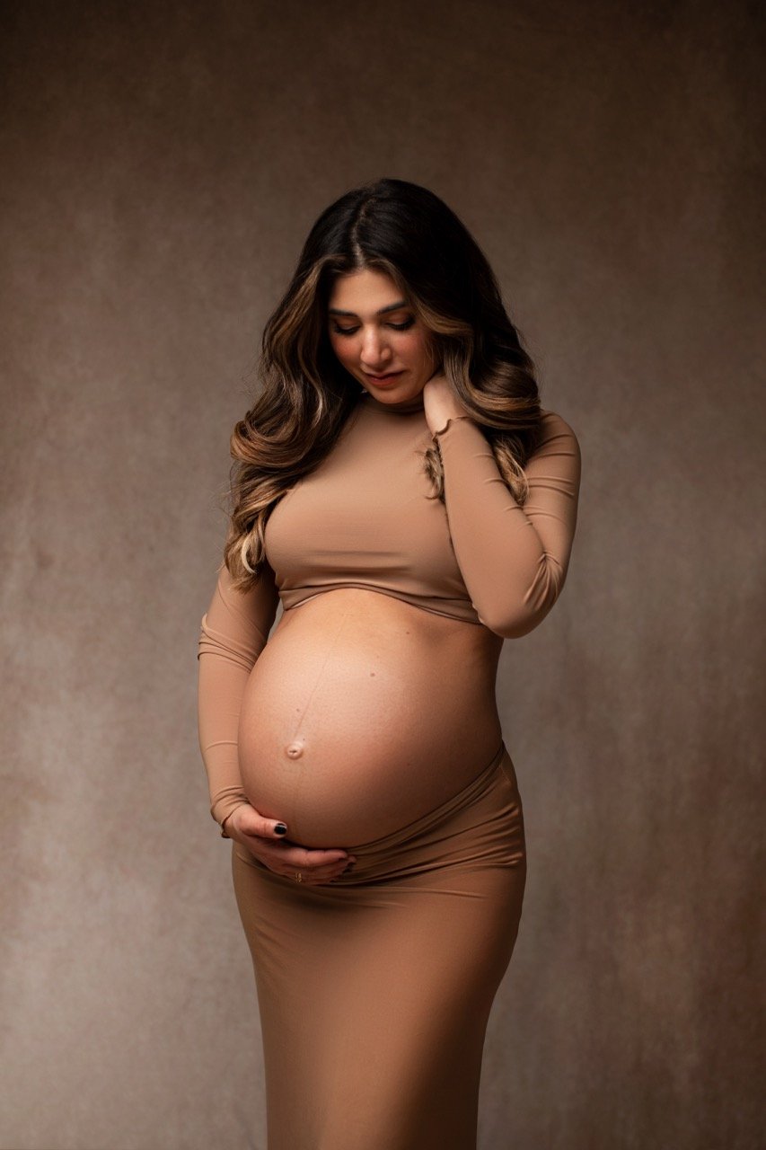 Schwangere Frau in hautengem, braunen Kleid hält ihren Babybauch, Hintergrund neutraler Farbton.