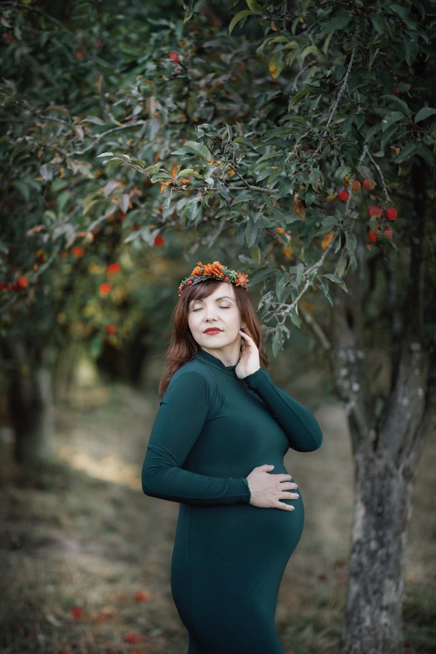 Schwangere Frau in grünem Kleid steht unter einem Baum, trägt Blütenkranz, Hand auf Bauch.