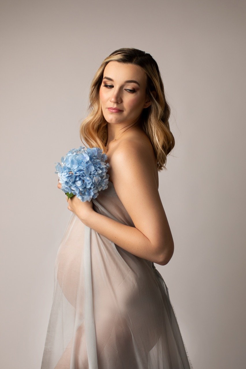 Schwangere Frau mit einem hellen Tuch bedeckt, hält einen Strauß blaue Blumen.