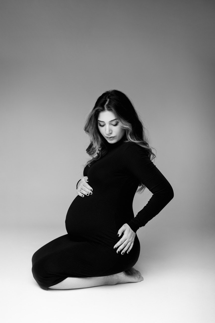 Schwarz-weiß Foto einer schwangeren Frau in einem schwarzen Kleid, die auf dem Boden kniet und ihre Hände auf ihrem Babybauch hält.
