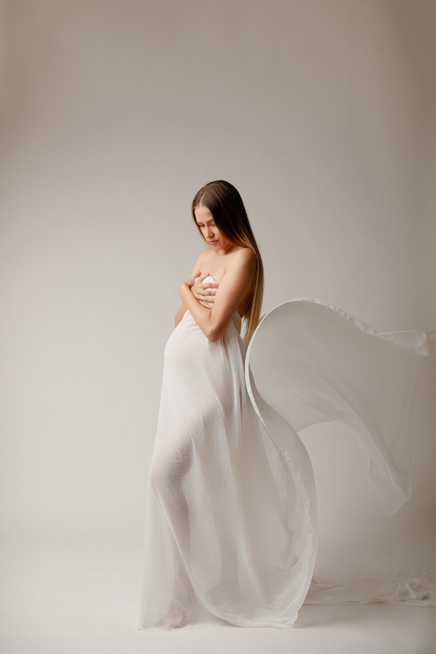 Eine schwangere Frau in einem fließenden weißen Kleid posiert vor einem neutralen Hintergrund.