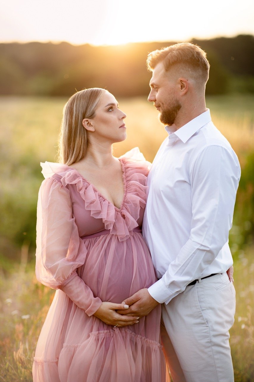 Schwangere Frau im rosa Kleid steht in einer sonnigen Wiese und wird von einem Mann in weißem Hemd liebevoll umarmt.