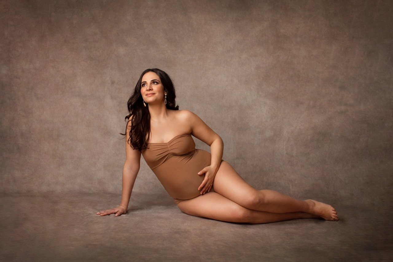 Schwangere Frau in einem beige Bodysuit sitzt auf dem Boden, Hintergrund in neutralen Tönen.