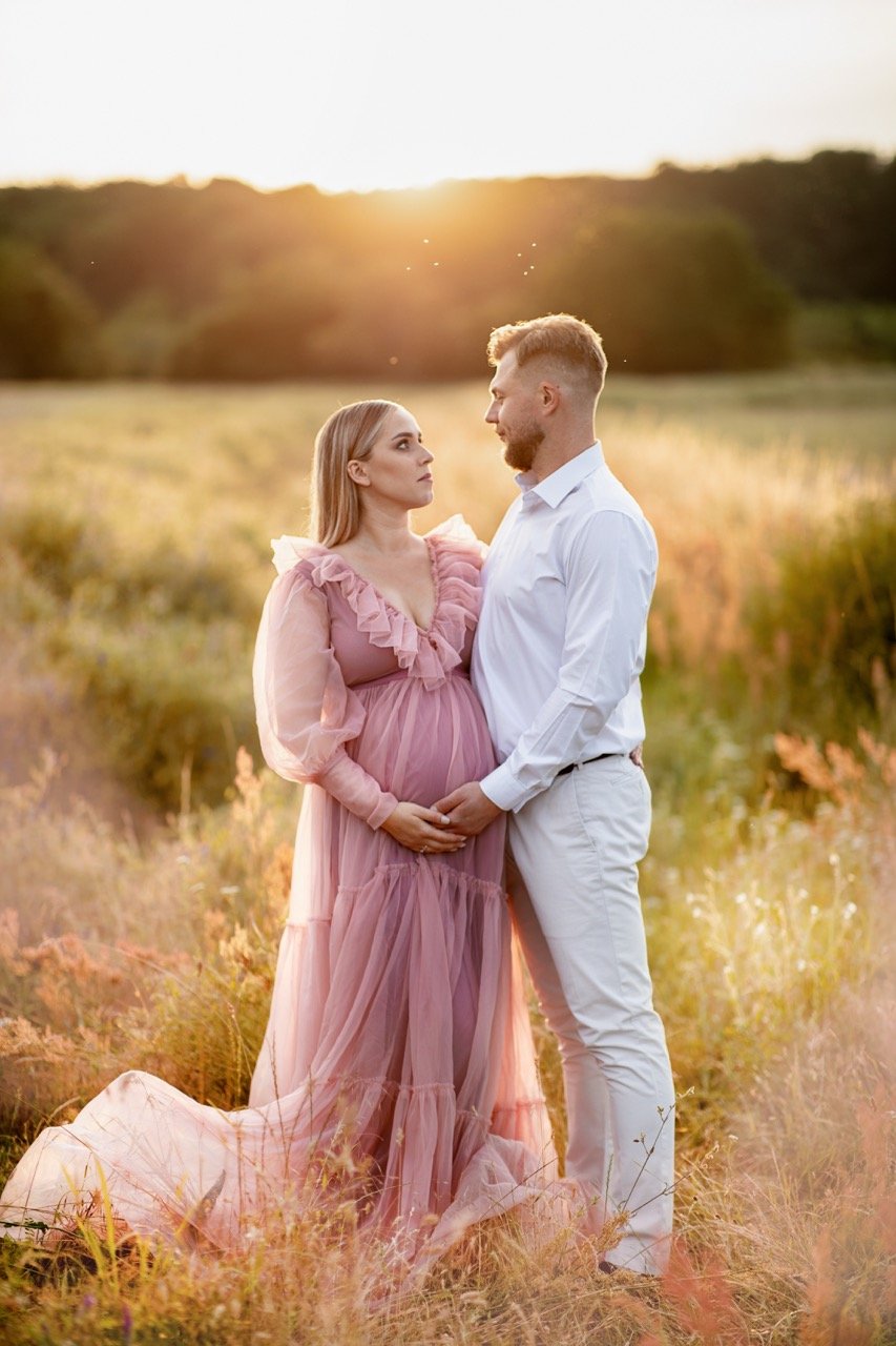 Schwangere Frau in rosa Kleid und Mann in weißem Hemd stehen in einem Feld und halten sich an den Händen, bei Sonnenuntergang.