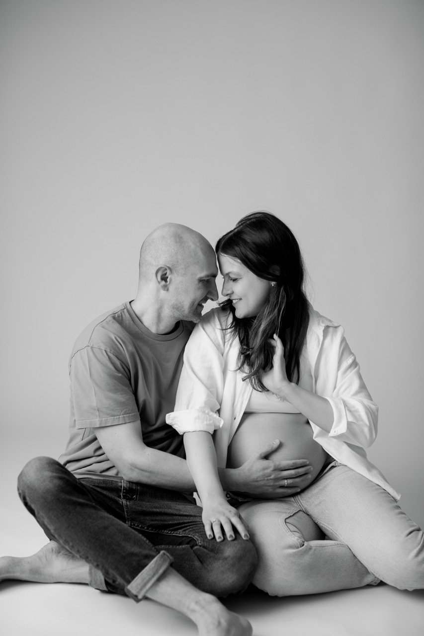 Schwarz-Weiß-Foto eines sitzenden Paares, das liebevoll aufeinander schaut. Der Mann legt seine Hand auf den Bauch der schwangeren Frau.
