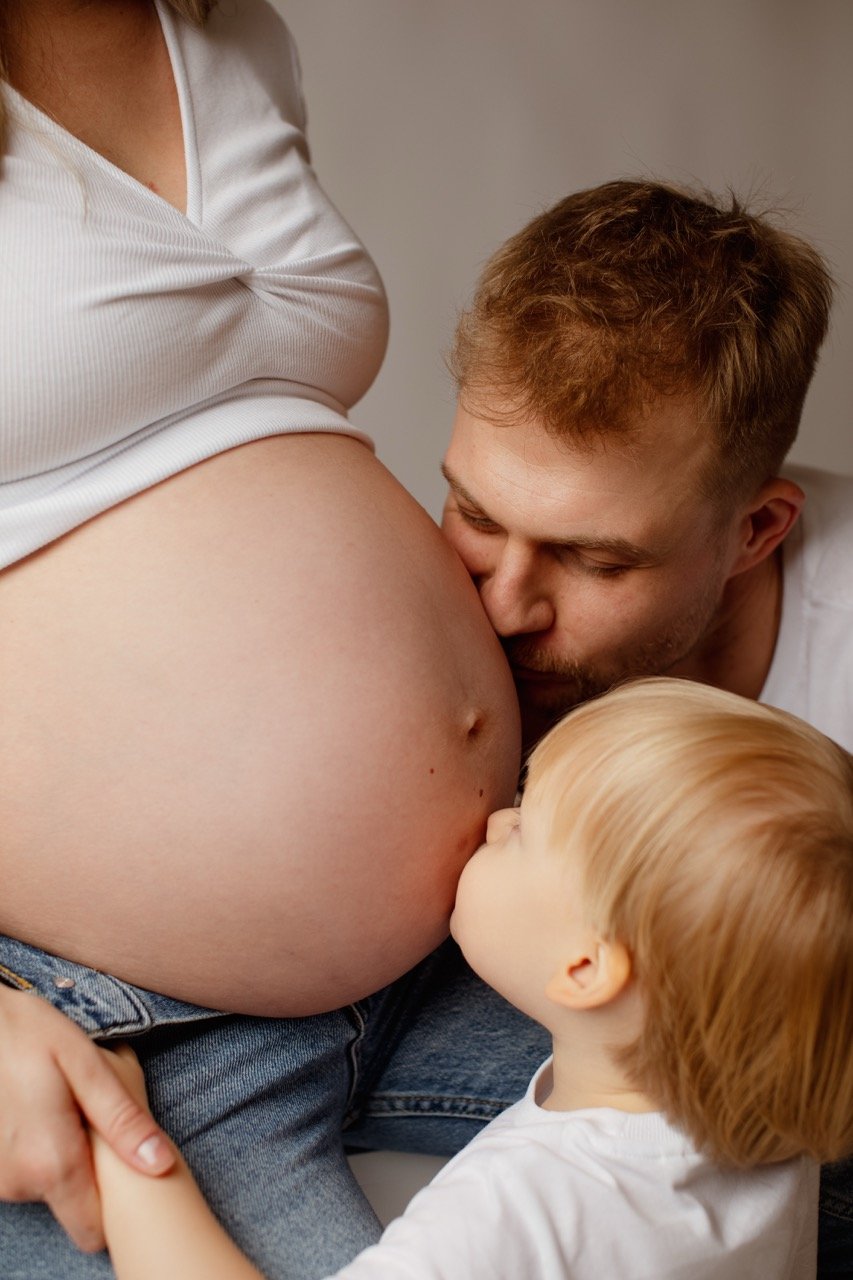 Ein Mann und ein kleines Kind küssen liebevoll den Babybauch einer schwangeren Frau.