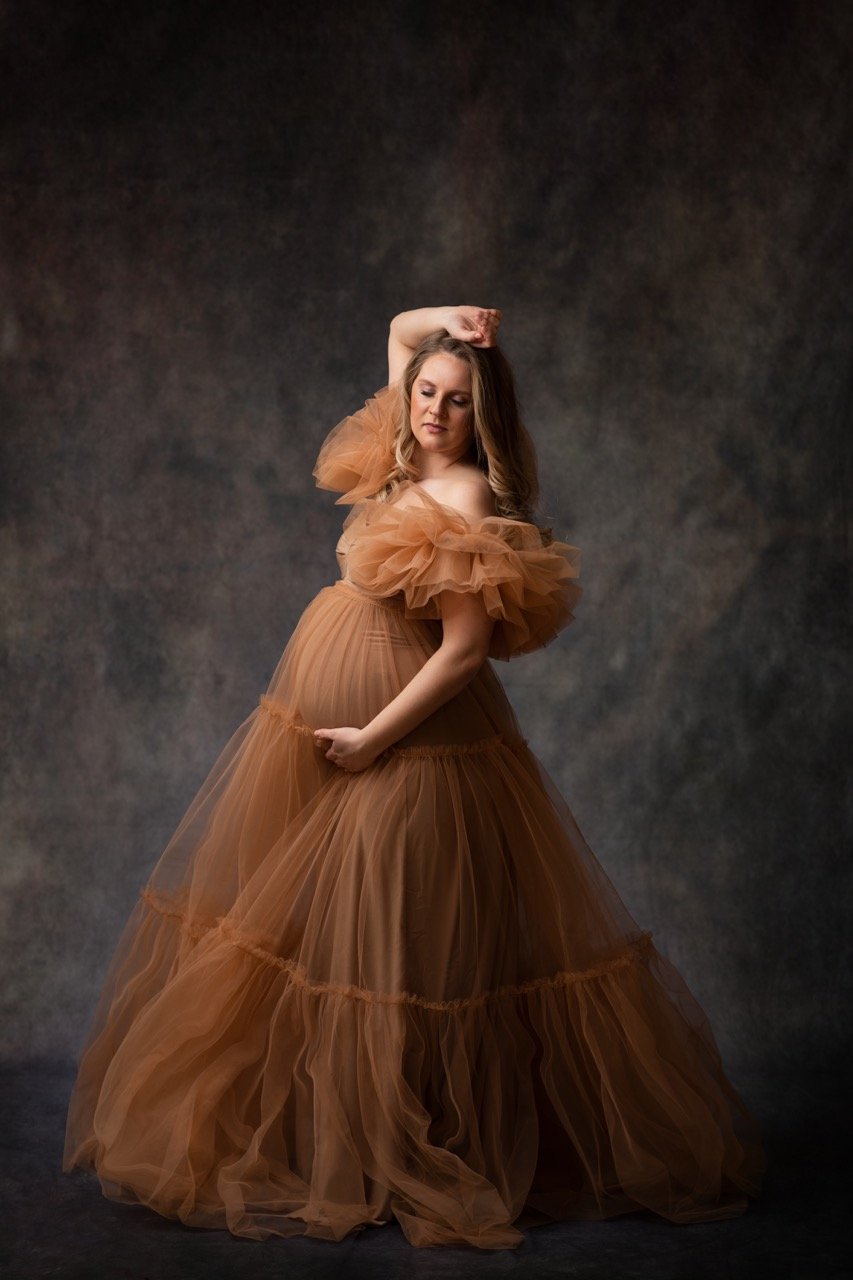 Eine schwangere Frau in einem langen, eleganten Kleid posiert vor einem dunklen Hintergrund. Sie hält ihren Bauch mit einer Hand und hat den anderen Arm über dem Kopf erhoben.