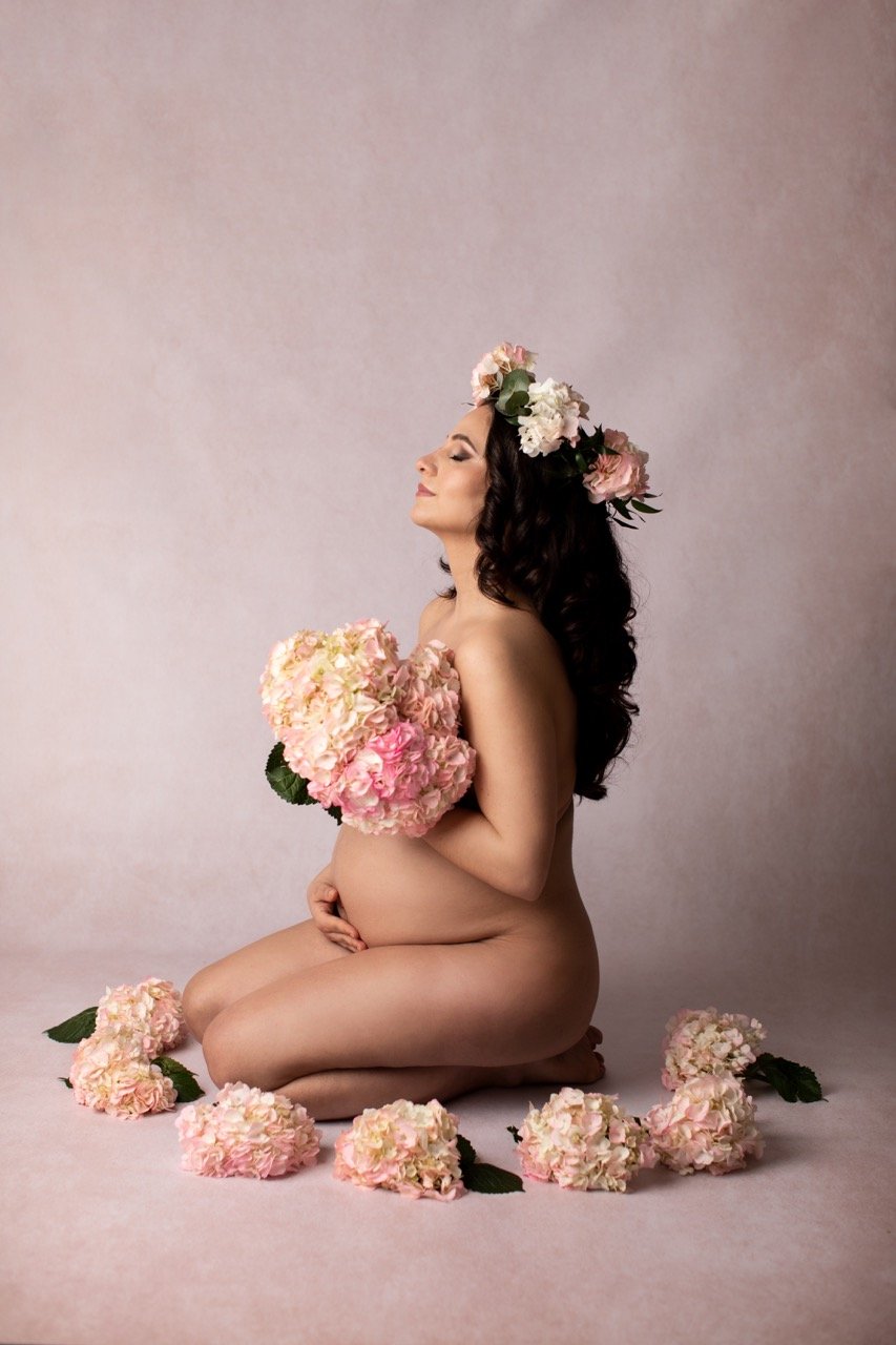 Eine schwangere Frau sitzt auf dem Boden mit geschlossenen Augen und hält einen Blumenstrauß aus rosa Hortensien. Sie trägt eine Blumenkrone und ist von weiteren Blumen umgeben. Der Hintergrund ist neutral.