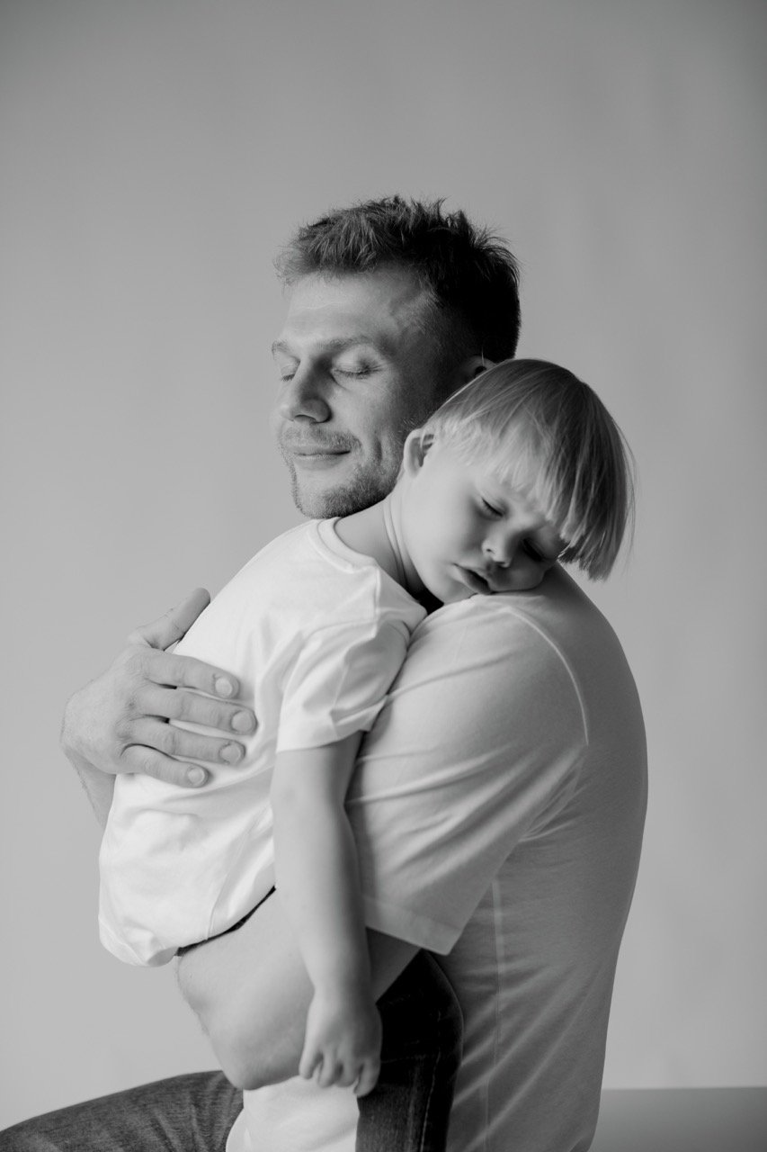 Ein Mann hält ein Kind in einem liebevollen Umarmung, beide haben die Augen geschlossen. Schwarz-Weiß-Foto.