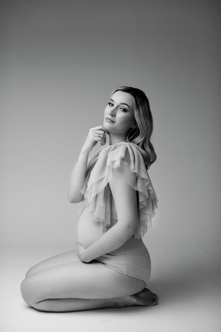 Schwarz-Weiß-Foto einer schwangeren Frau, die kniend posiert. Sie trägt ein durchsichtiges Oberteil und hält sich am Bauch. Der Hintergrund ist neutral.