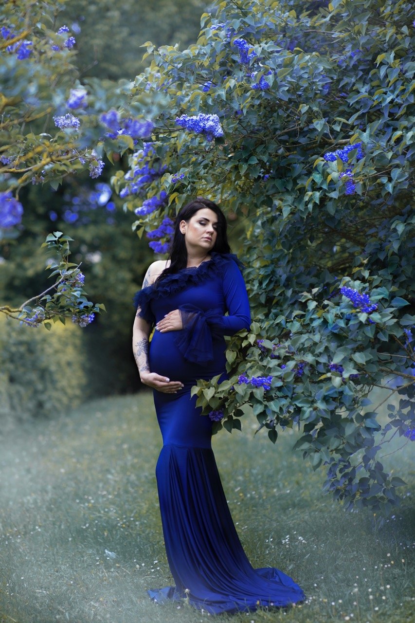 Schwangere Frau in blauem Kleid steht in einem Garten mit lila blühenden Bäumen.