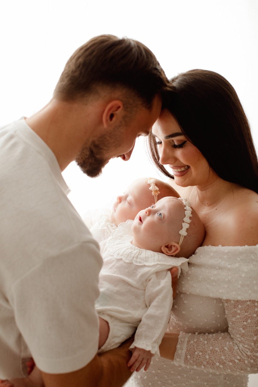 Eine lächelnde Frau und ein Mann schauen auf zwei Babys in weißen Kleidern. Die Babys tragen Blumenhaarbänder. Die Szene strahlt Wärme und Liebe aus.