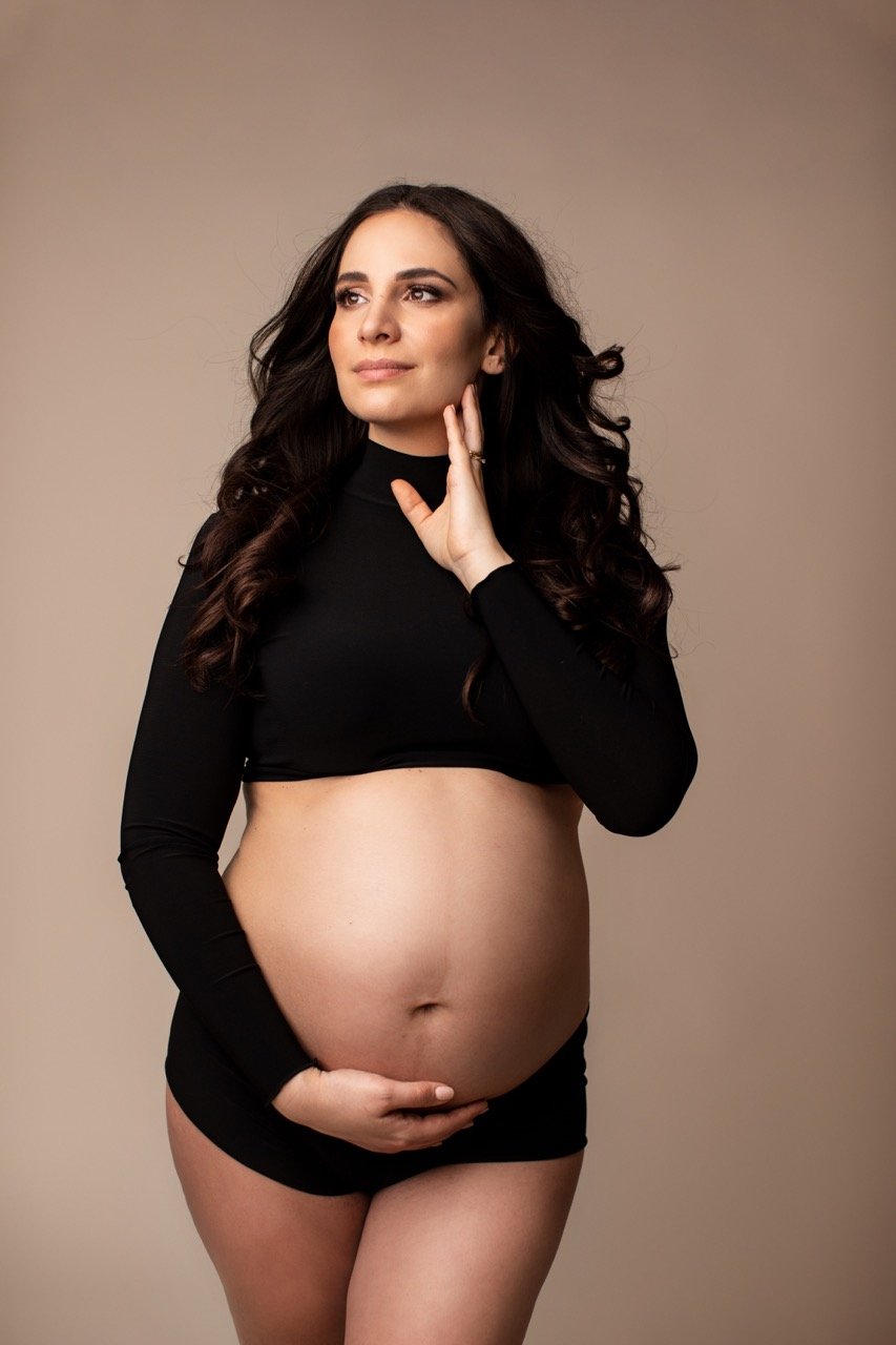 Schwangere Frau in schwarzem Outfit, die ihren Bauch hält, vor neutralem Hintergrund.
