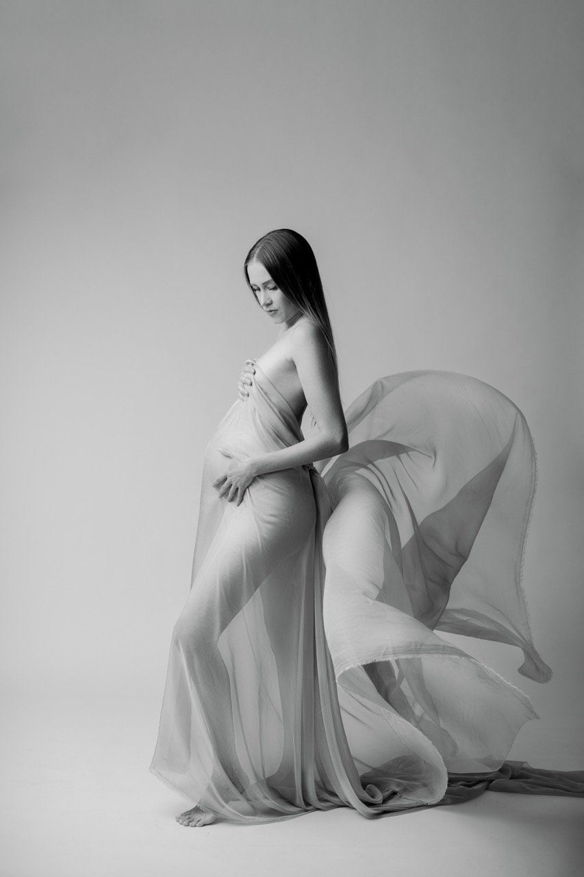 Schwarzweiß-Fotografie einer schwangeren Frau, die in transparente Stoffe gehüllt ist, mit fließenden Linien, die Bewegung andeuten.