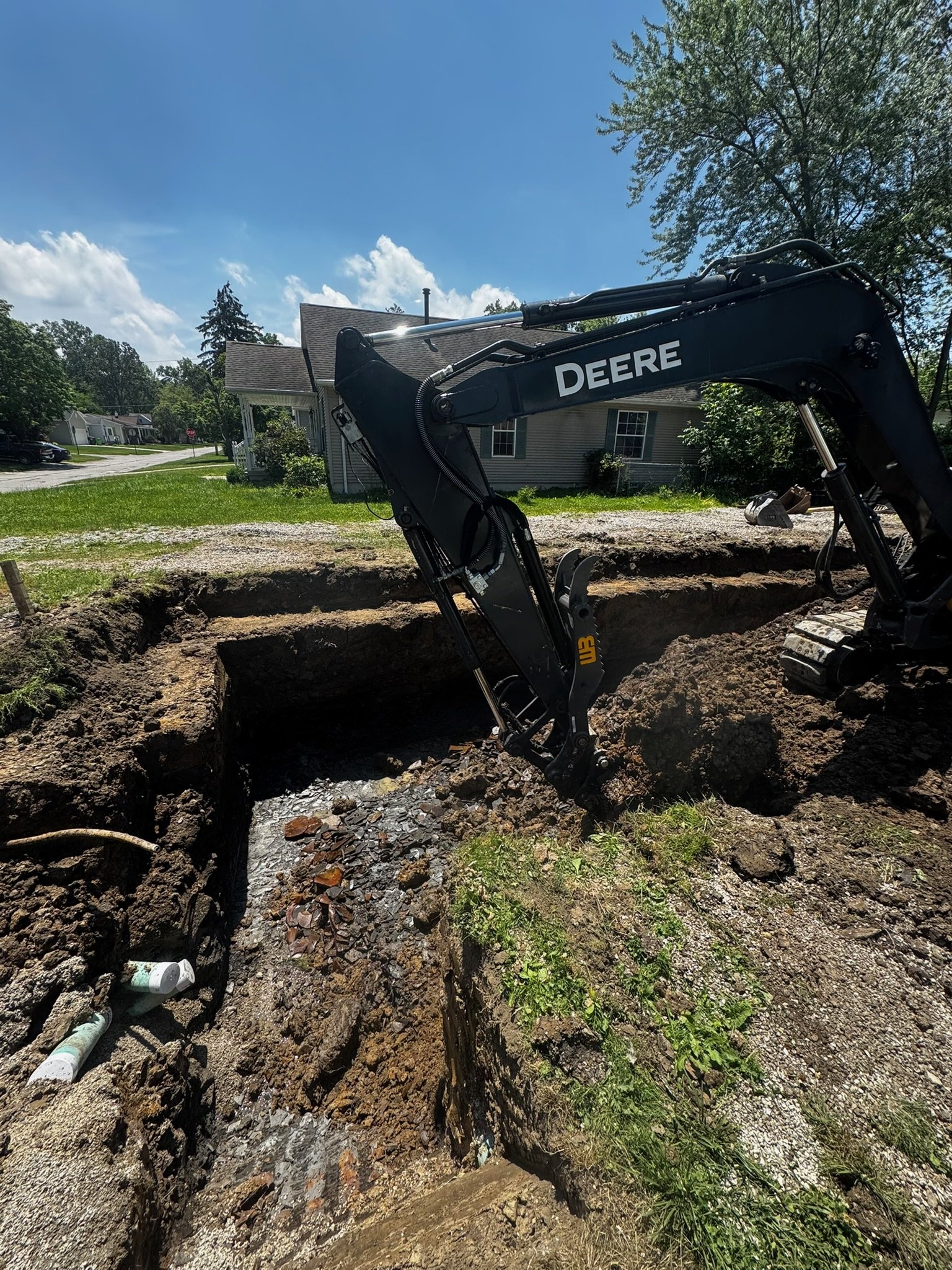 A construction site with an excavator digging a trench in a residential area with houses, trees, and a partly cloudy sky.