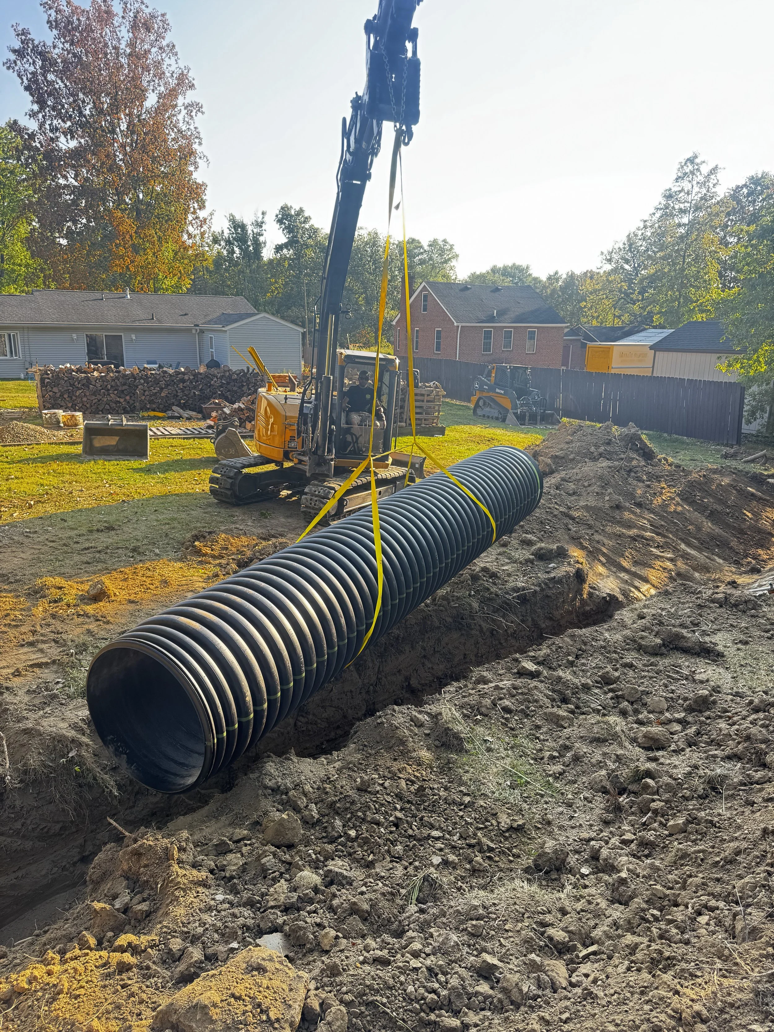 A large black corrugated pipe being lowered into a trench by a crane on a construction site in a backyard, with a small excavator nearby and residential houses in the background.