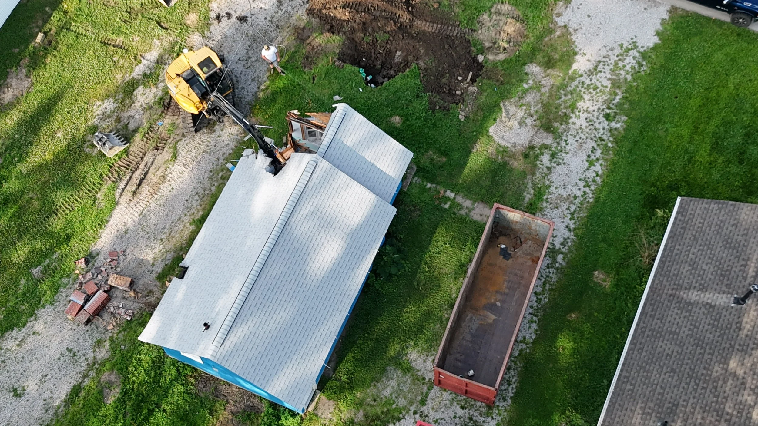 Aerial view of a construction site with a house, a drop-off container, and workers. A crane is removing part of the roof, and a person is standing nearby. The yard has grass, gravel, and some construction debris.