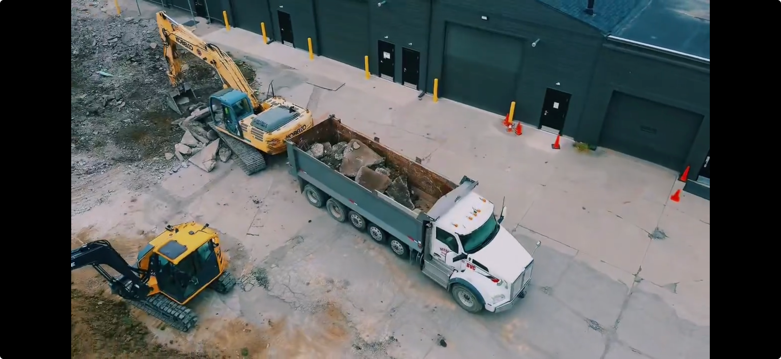 Construction site with a white dump truck carrying rocks, a yellow excavator digging and loading dirt, and a small yellow excavator on the ground. The background is a dark green building with orange cones and yellow safety posts along the sidewalk.