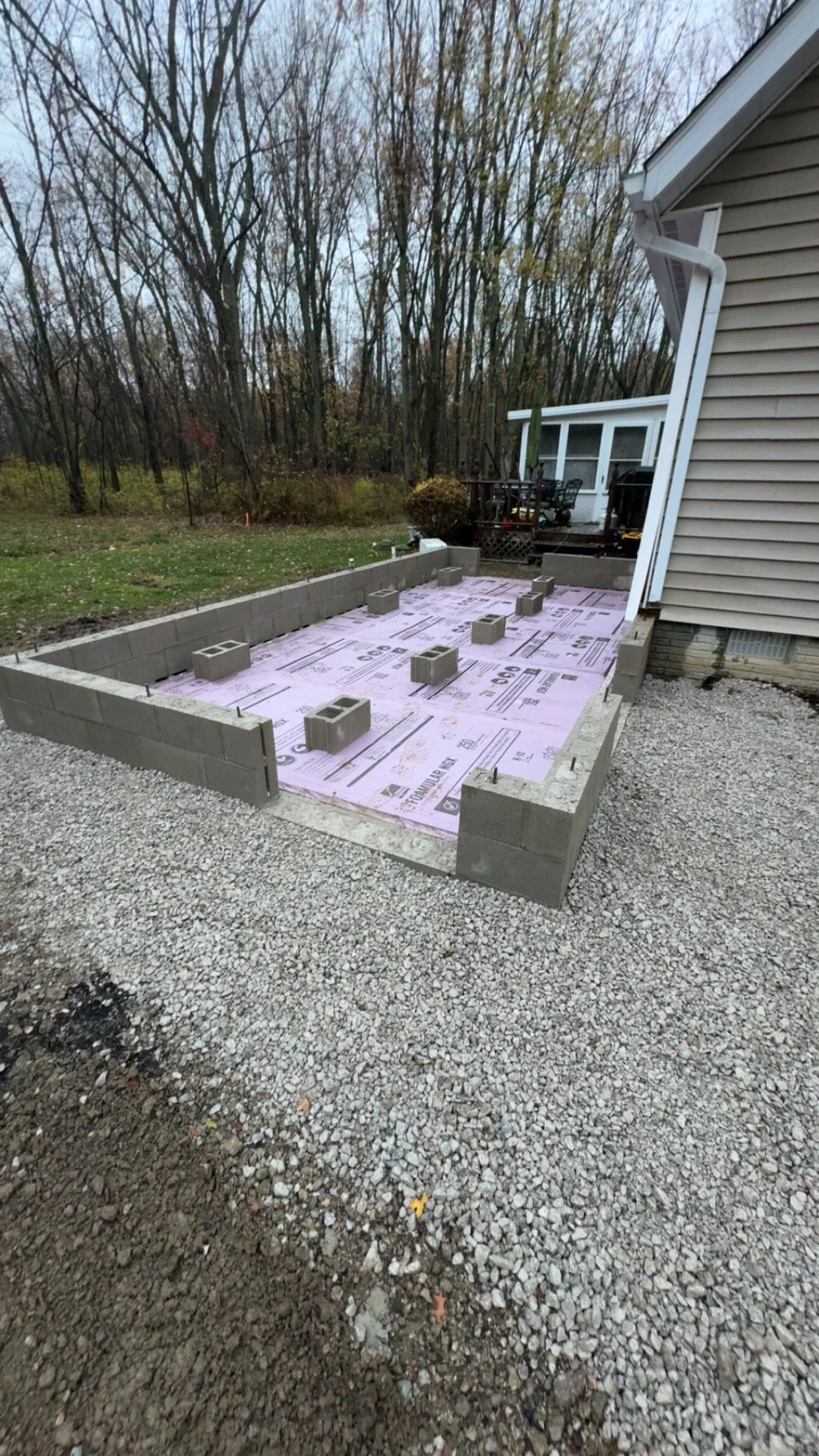 Construction site of a small building or extension with cinder blocks and pink weatherproofing material, adjacent to a house with beige vinyl siding, in a backyard with leafless trees.