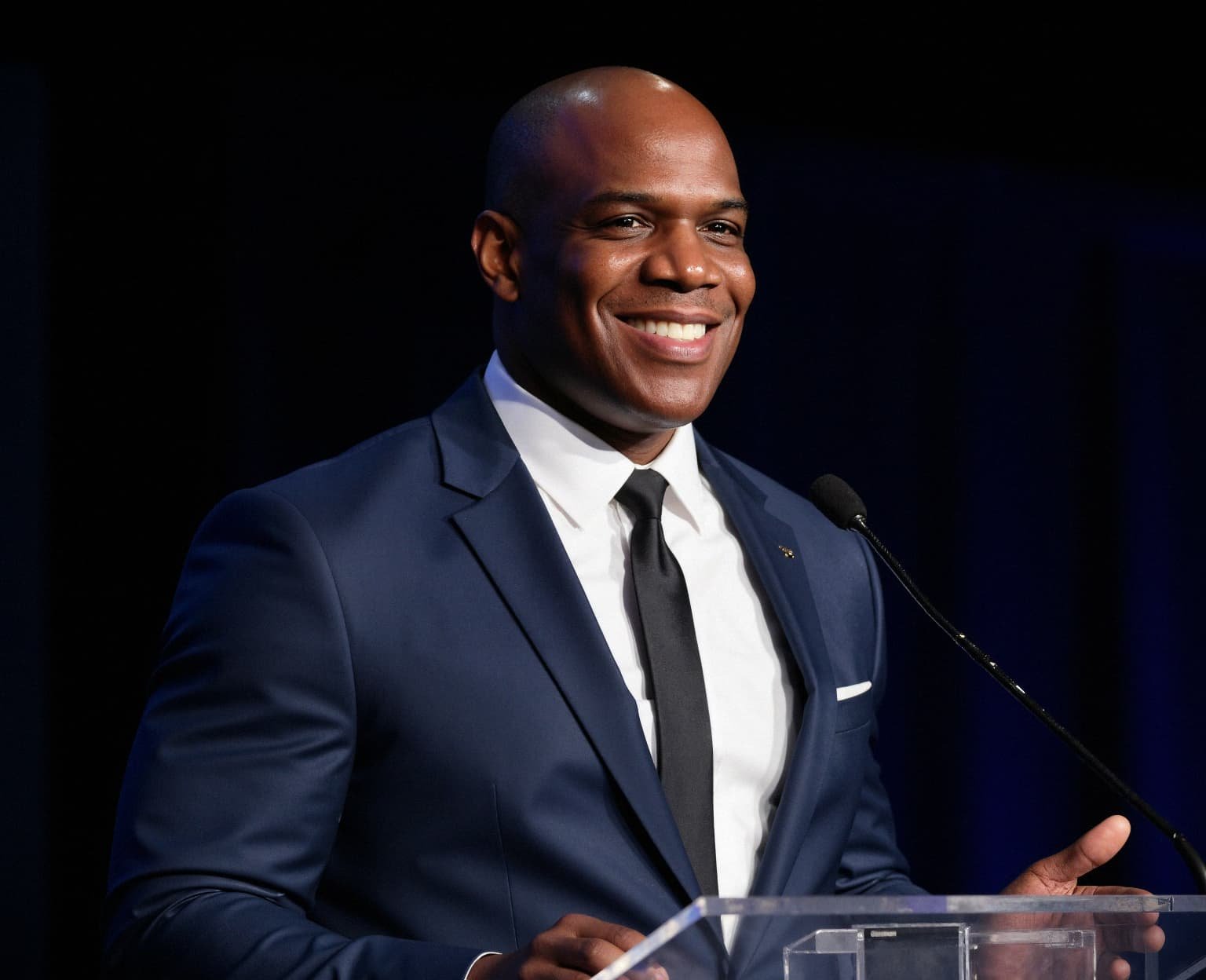 A smiling man in a navy suit, white shirt, and black tie standing at a podium with a microphone.