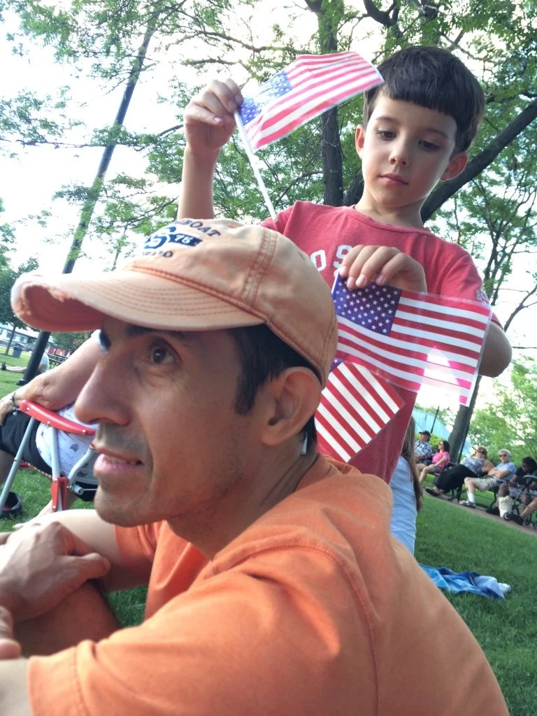 A man in an orange shirt and baseball cap is lying on the grass, with a young boy standing behind him. The boy is holding and waving small American flags. They are outdoors on a sunny day with trees and other people sitting on benches in the backgrou