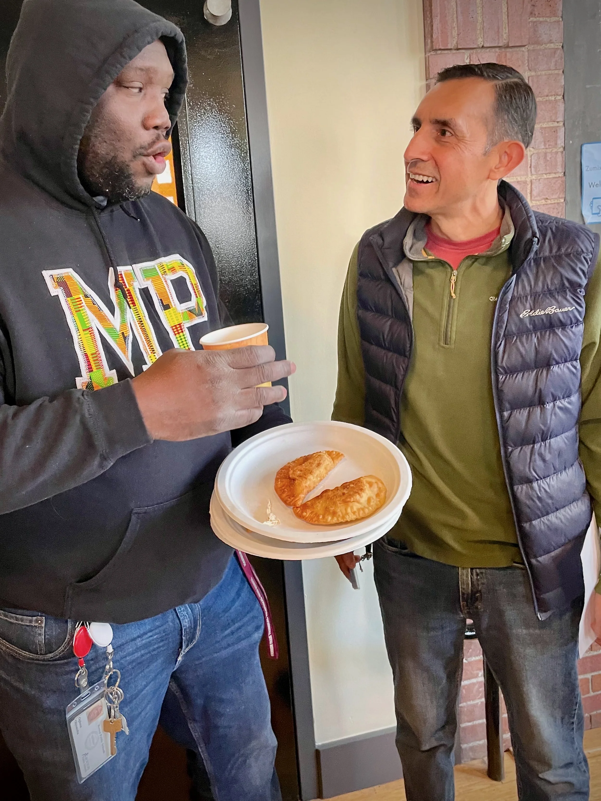 Two men are engaged in conversation inside a restaurant. One man is holding a paper plate with two fried empanadas and a cup. The man on the left is wearing a black hoodie with colorful lettering, and the man on the right is wearing a green shirt wit