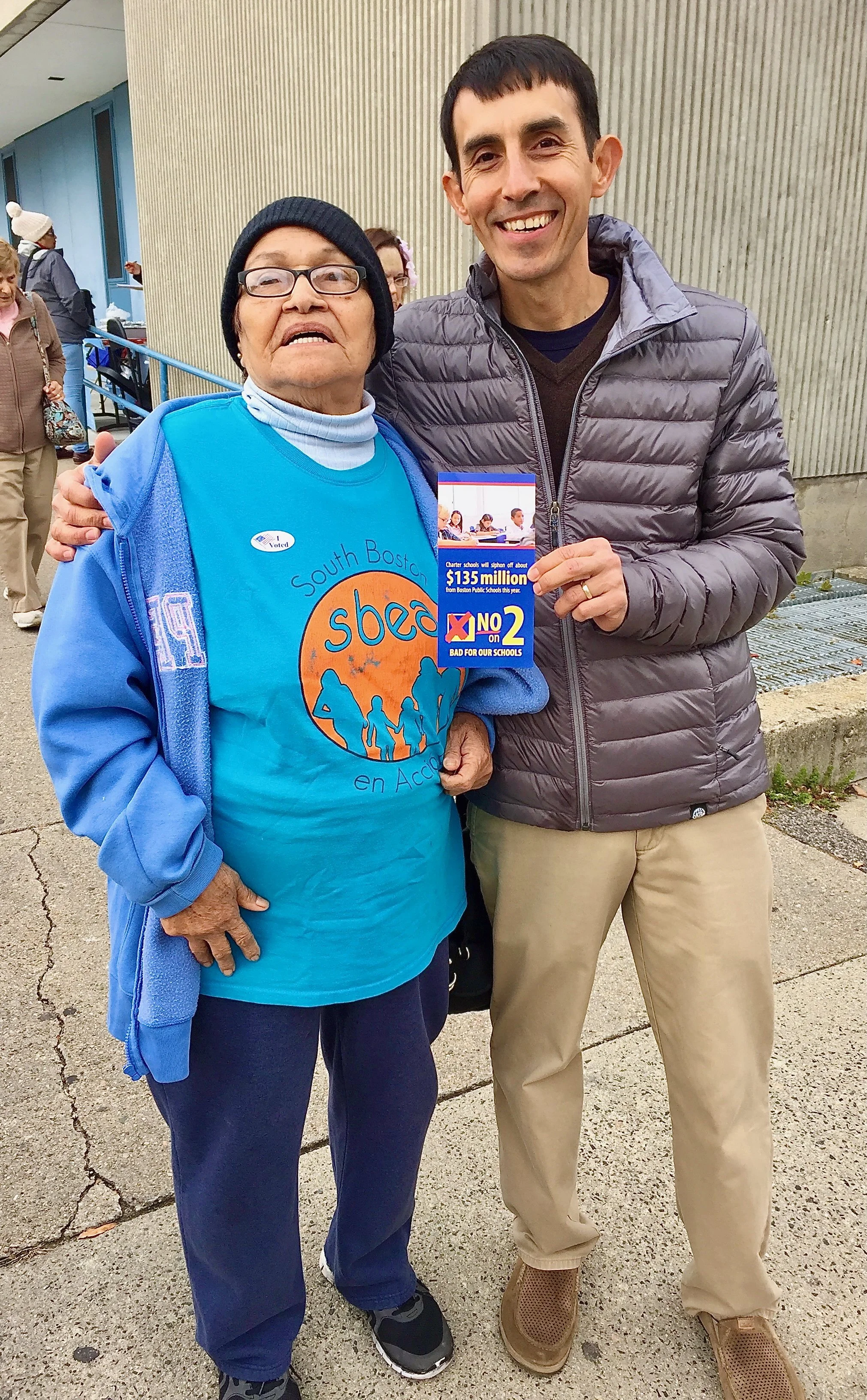 A woman wearing a blue t-shirt and jacket standing next to a man in a gray jacket holding a campaign sign, both smiling outdoors near a building.