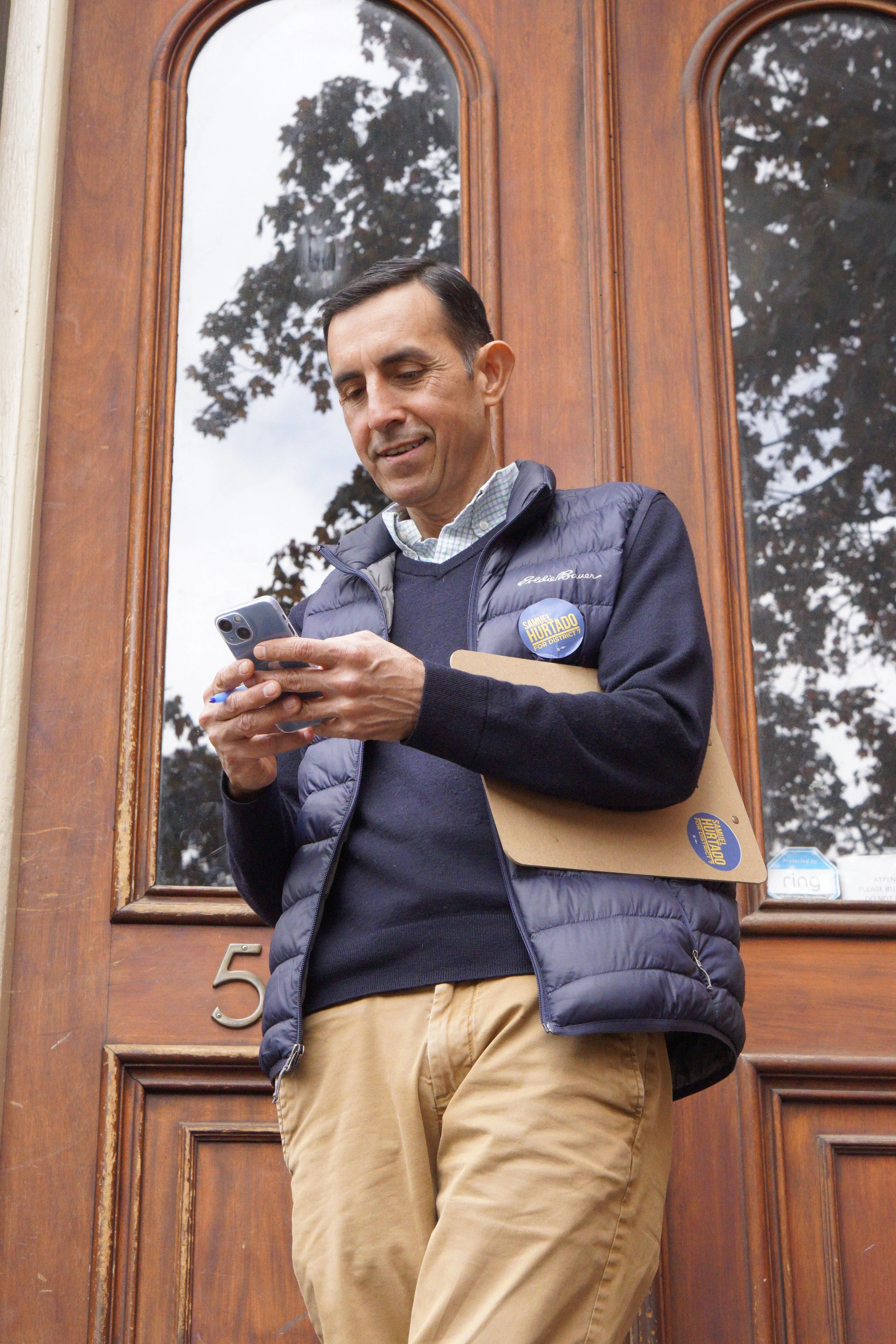 Samuel Hurtado stands outside while canvassing, looking at his phone, holding a folder, wearing a campaign sticker and a navy vest.