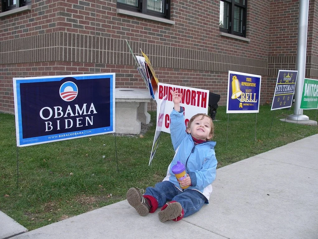 A young child sitting on the sidewalk holding a flag with campaign signs from various political candidates and campaigns behind him, including Obama Biden and others.