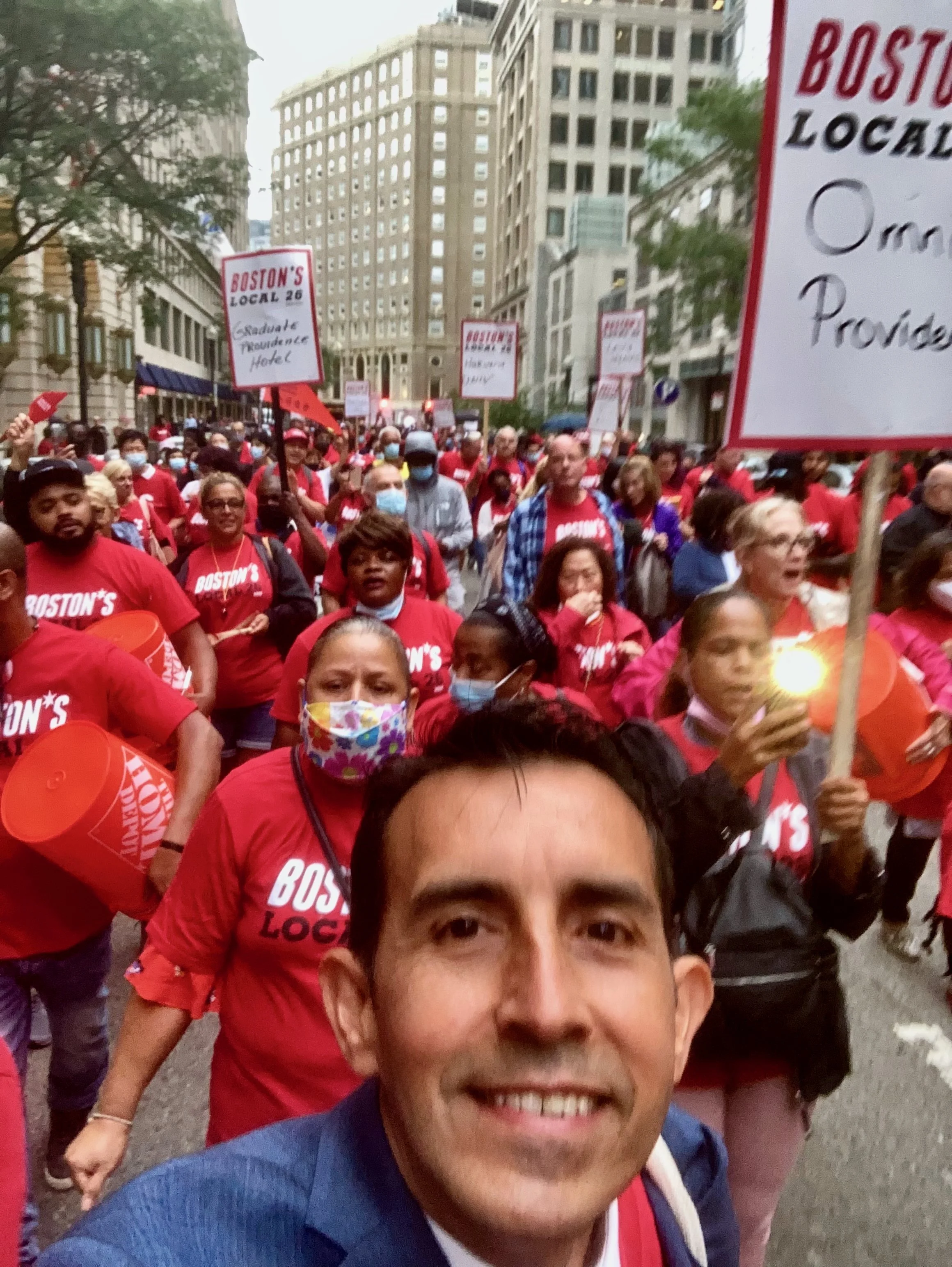 A man taking a selfie at a protest march with a crowd of people behind him wearing red shirts and holding signs, on a city street with tall buildings.