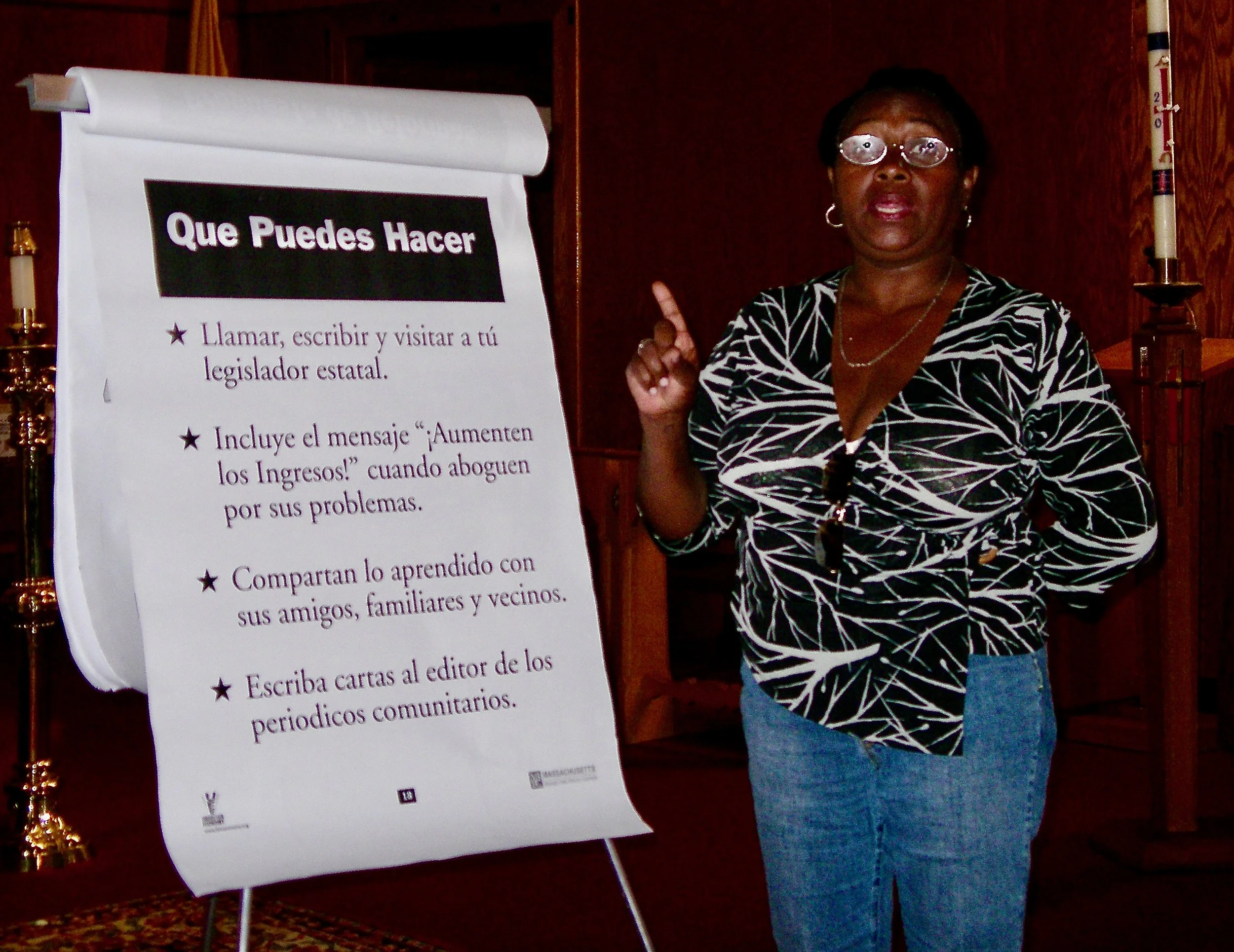 A woman standing next to a large presentation board with a list in Spanish titled 'Que Puedes Hacer.' She is wearing glasses, a black and white patterned blouse, and blue jeans, and is pointing at the board with her index finger.