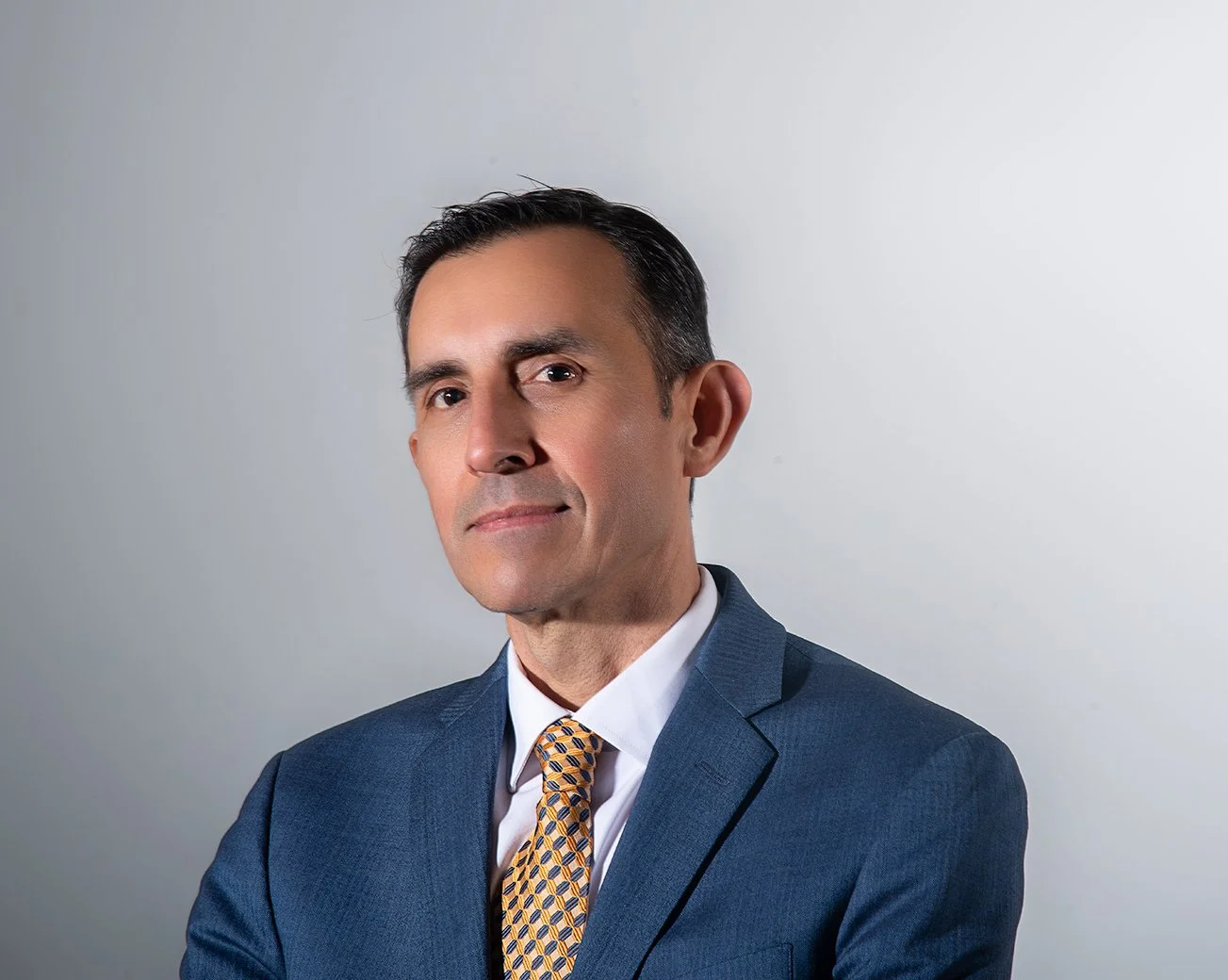 Portrait of a Samuel Hurtado in a blue suit with a patterned yellow tie, looking directly at the camera against a light gray background.