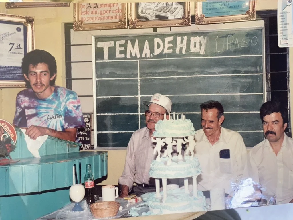 A birthday celebration with three men sitting in front of a large blue birthday cake decorated with white and blue icing, and a smaller cake on top. A young man in a tie-dye shirt stands to the left, smiling. The background features a chalkboard with