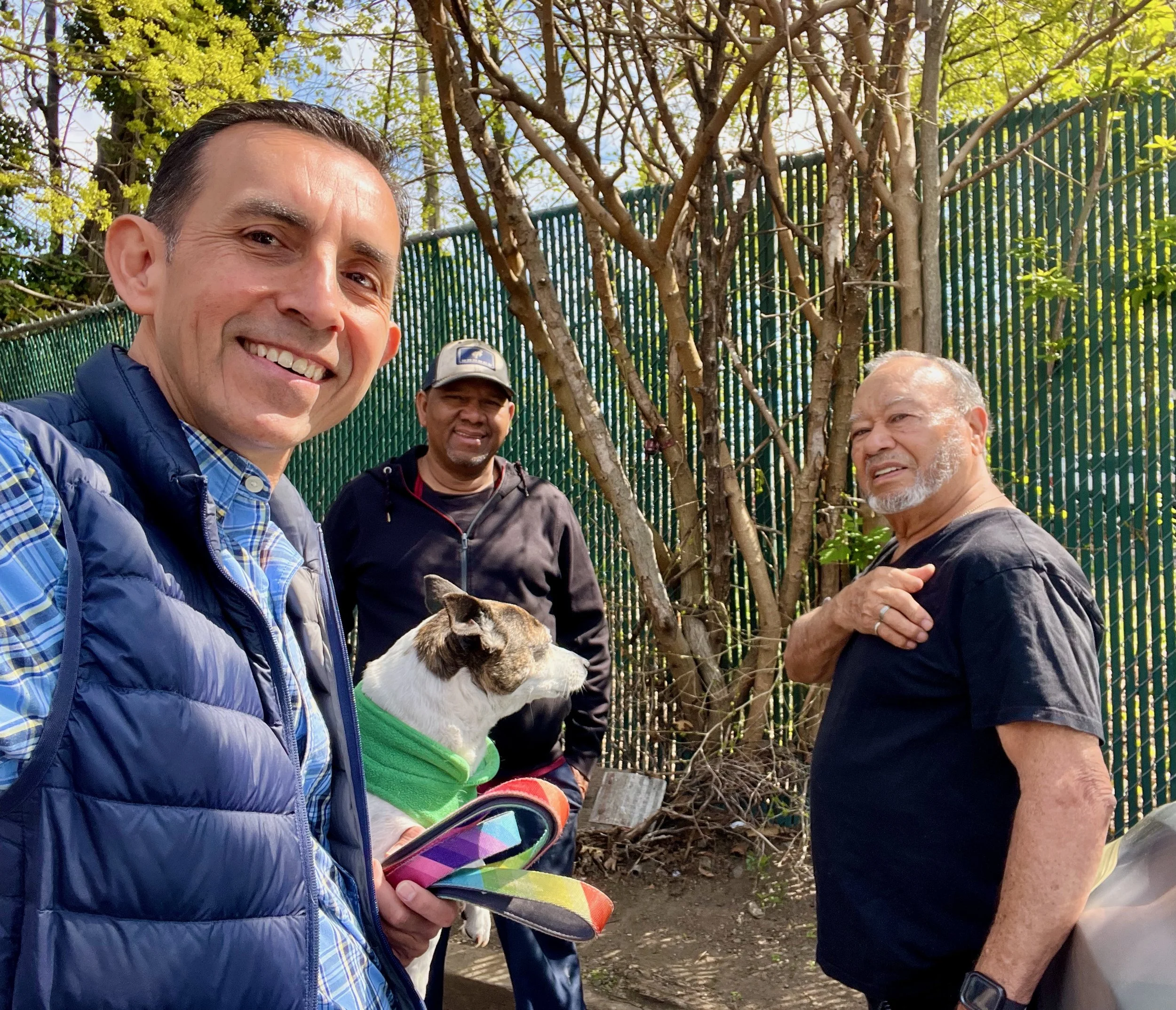 Three men and a dog outdoors near a green fence with trees in the background. One man is smiling at the camera, holding a colorful tablet, and wearing a blue vest and plaid shirt. The second man is wearing a dark jacket and a gray cap, and the third