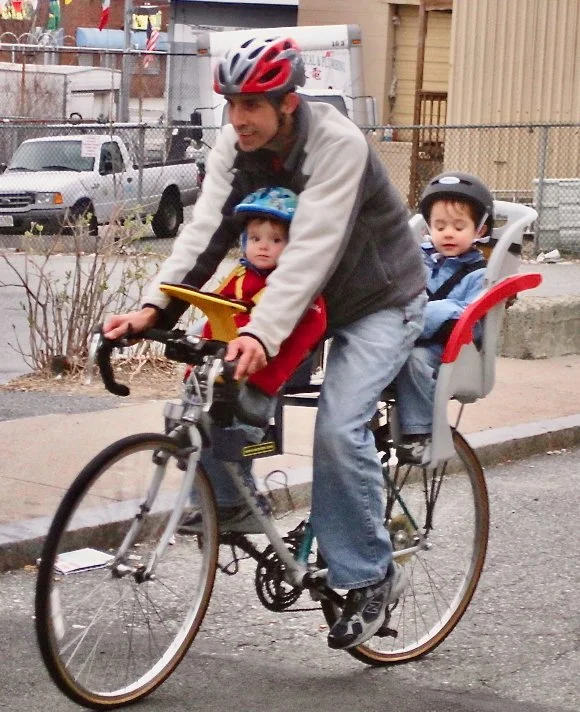 A man and two children riding a bicycle on a sidewalk, with the two children seated in a child bike seat, all wearing helmets.