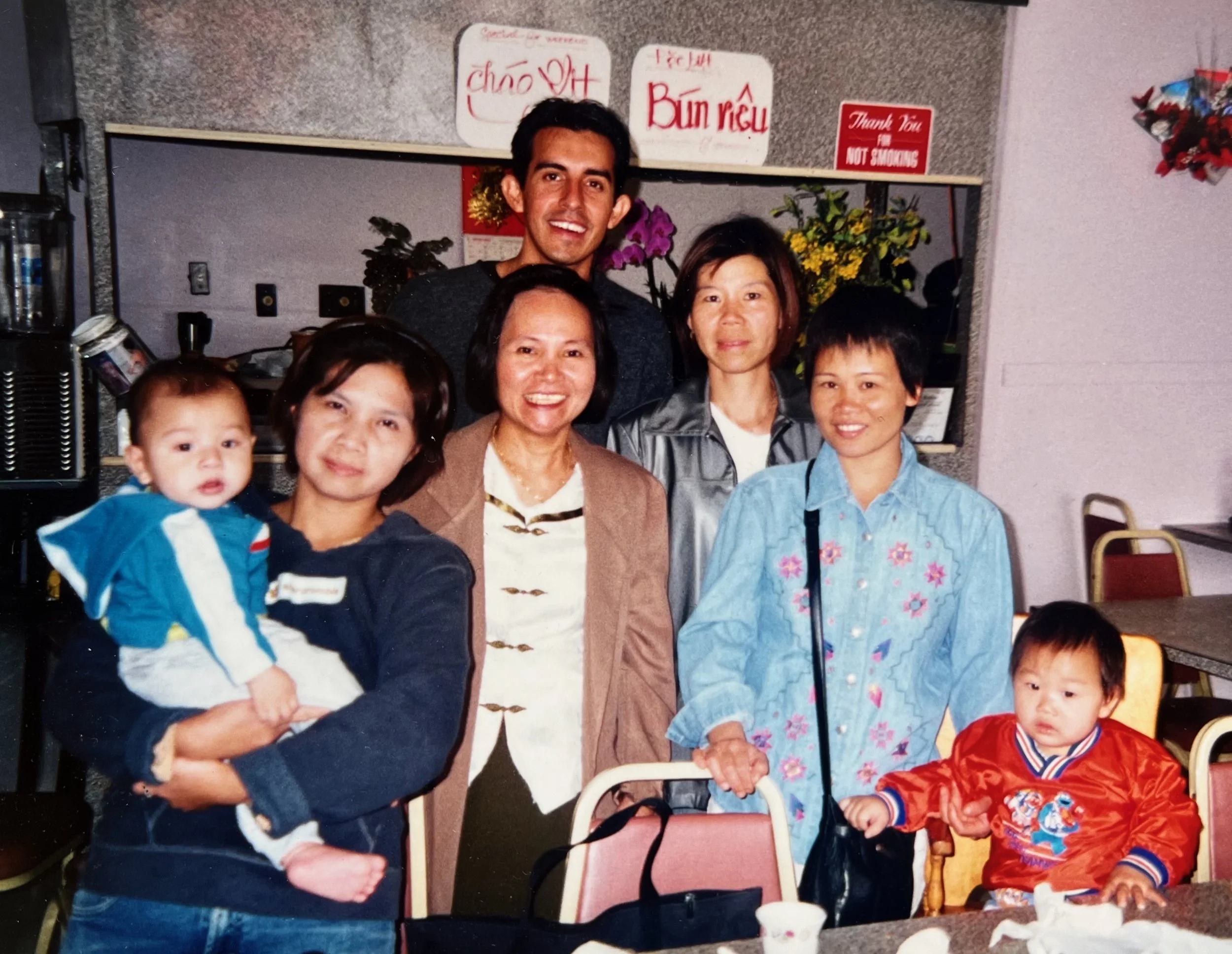 Group of seven people, including two children, posing together in a restaurant or banquet hall. Two women and one man standing behind, two children seated in front, one being held by an adult. The background shows flowers, signs in Vietnamese, and a