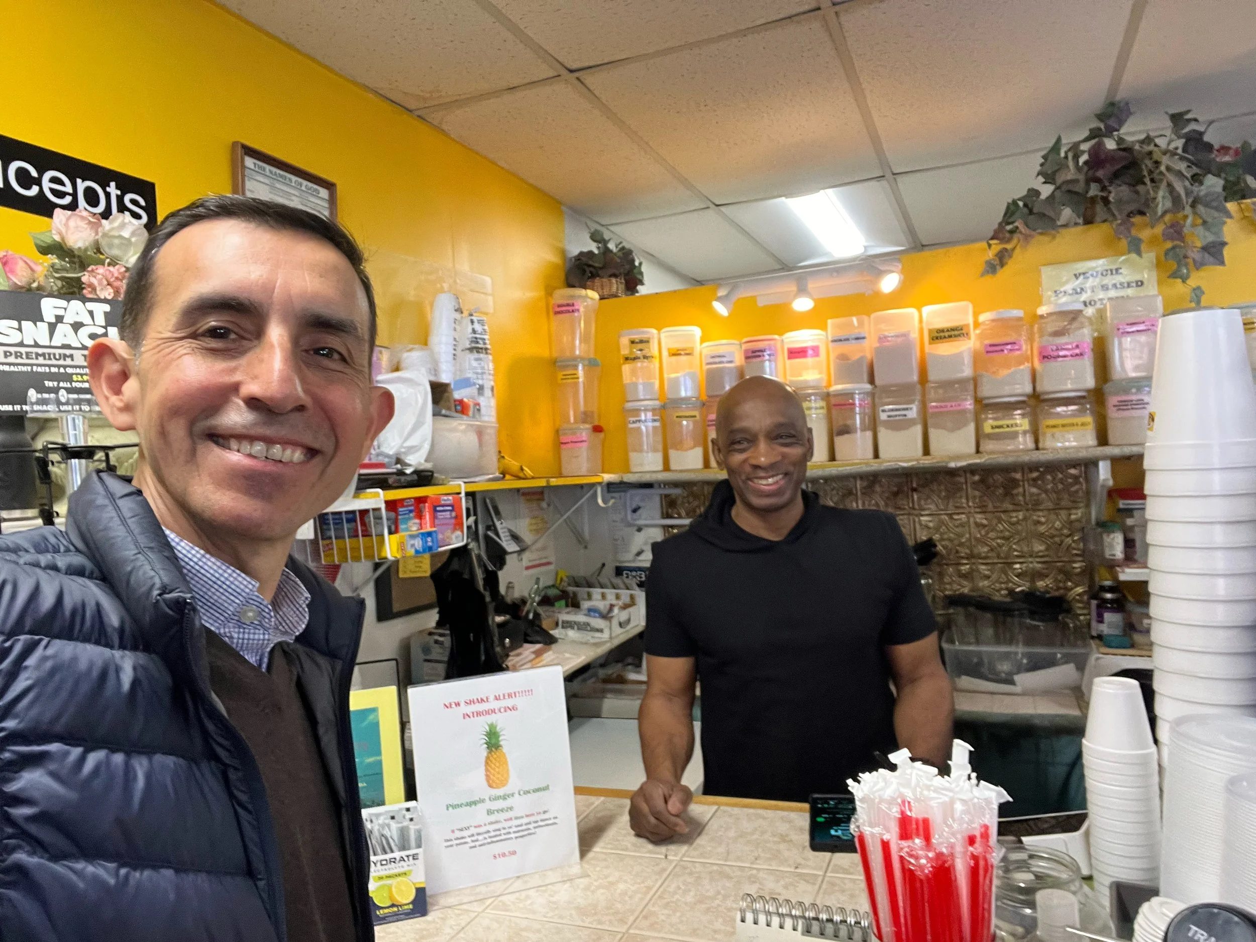 Two men smiling inside a colorful drink shop with containers of powders on the shelves behind them and a counter with cups and utensils.