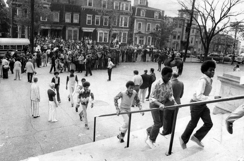 A large group of people, including children and adults, gathered outside a Boston school, some climbing stairs to enter while others standing or walking in the open area.
