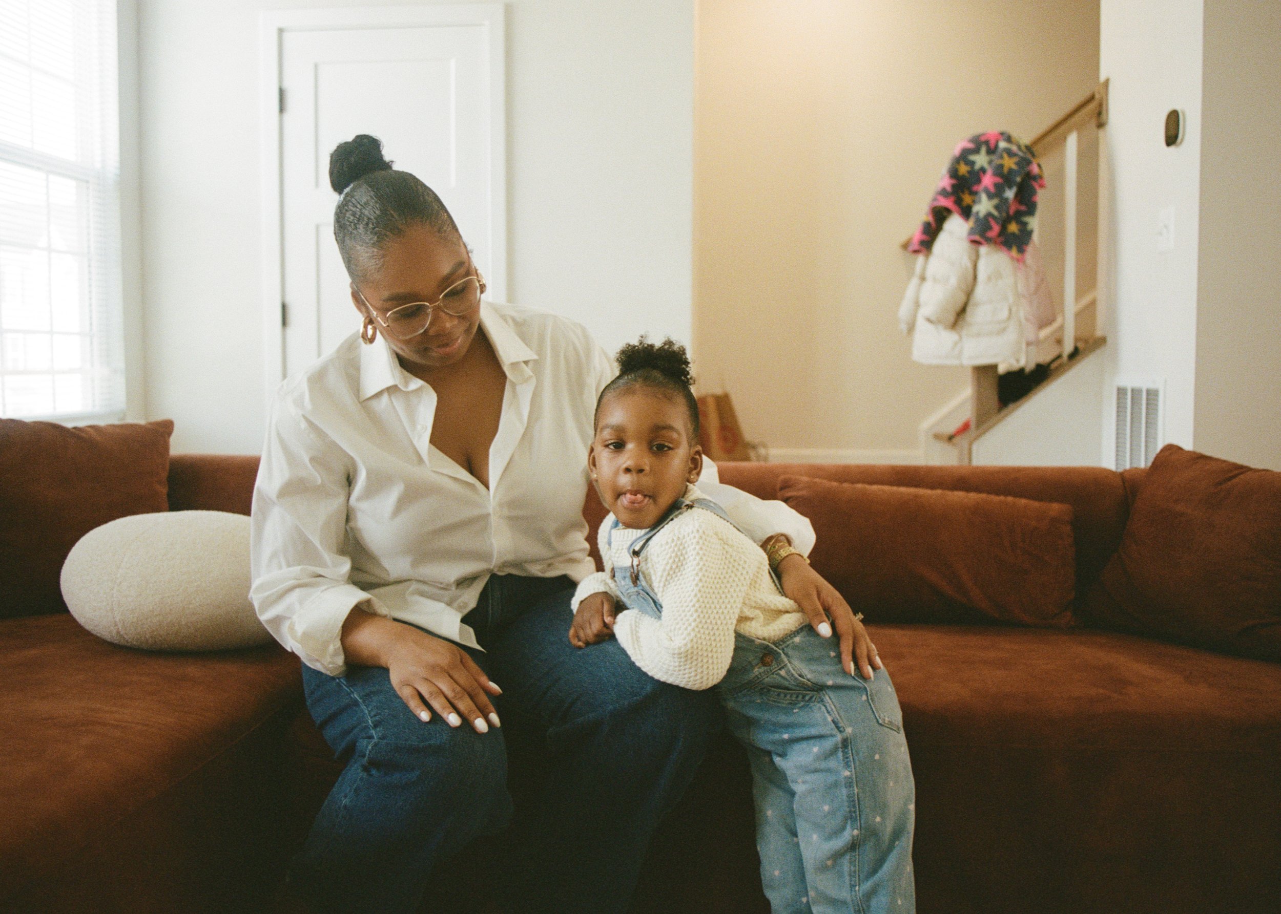 A woman sitting on a brown couch with a child, both indoors in front of a window. The woman wears glasses and a white shirt, while the child wears a white sweater and denim overalls. Coats hang on a railing in the background.