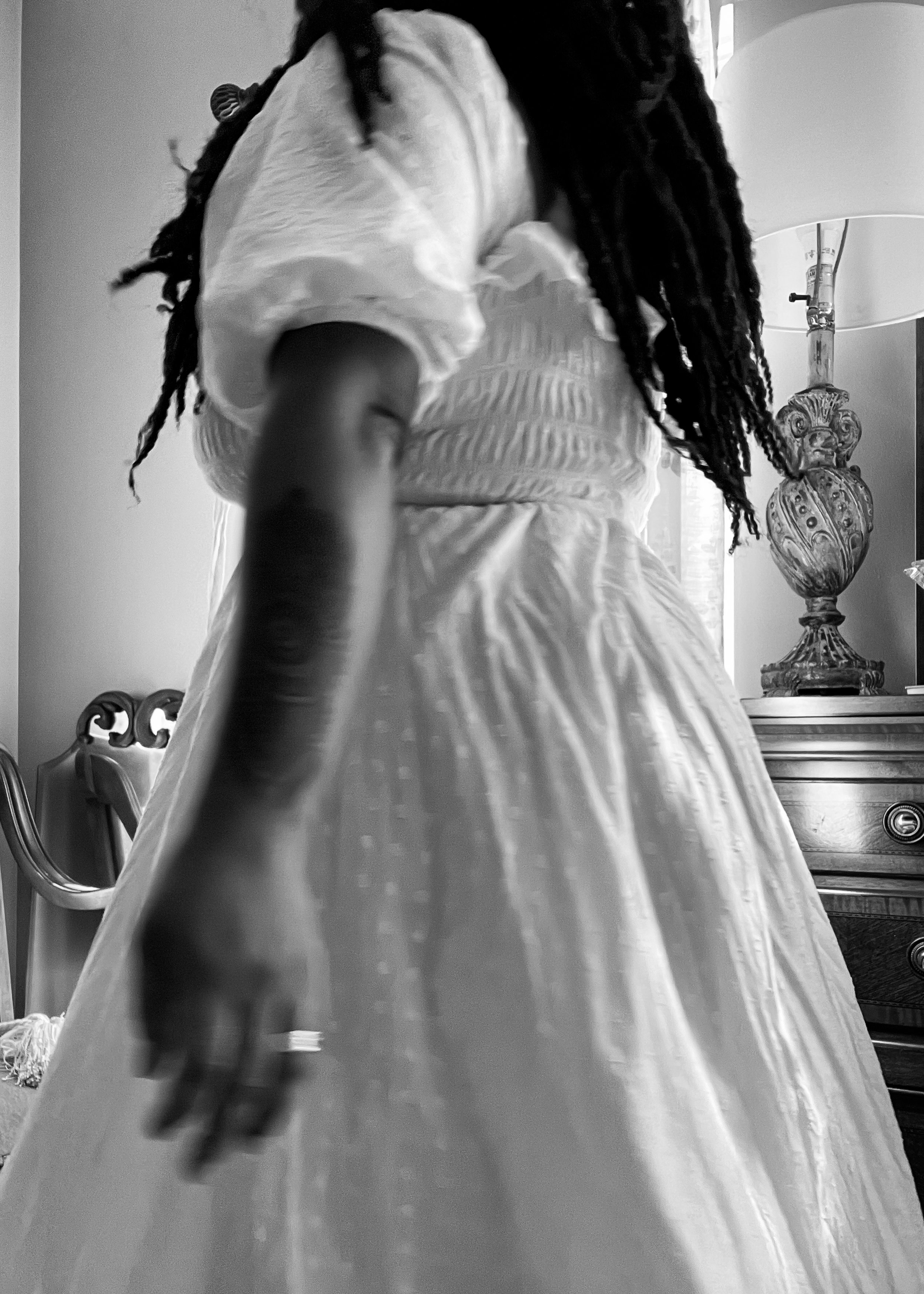 Black and white image of a person in a white dress, standing in a room with a chair, lamp, and ornate table.