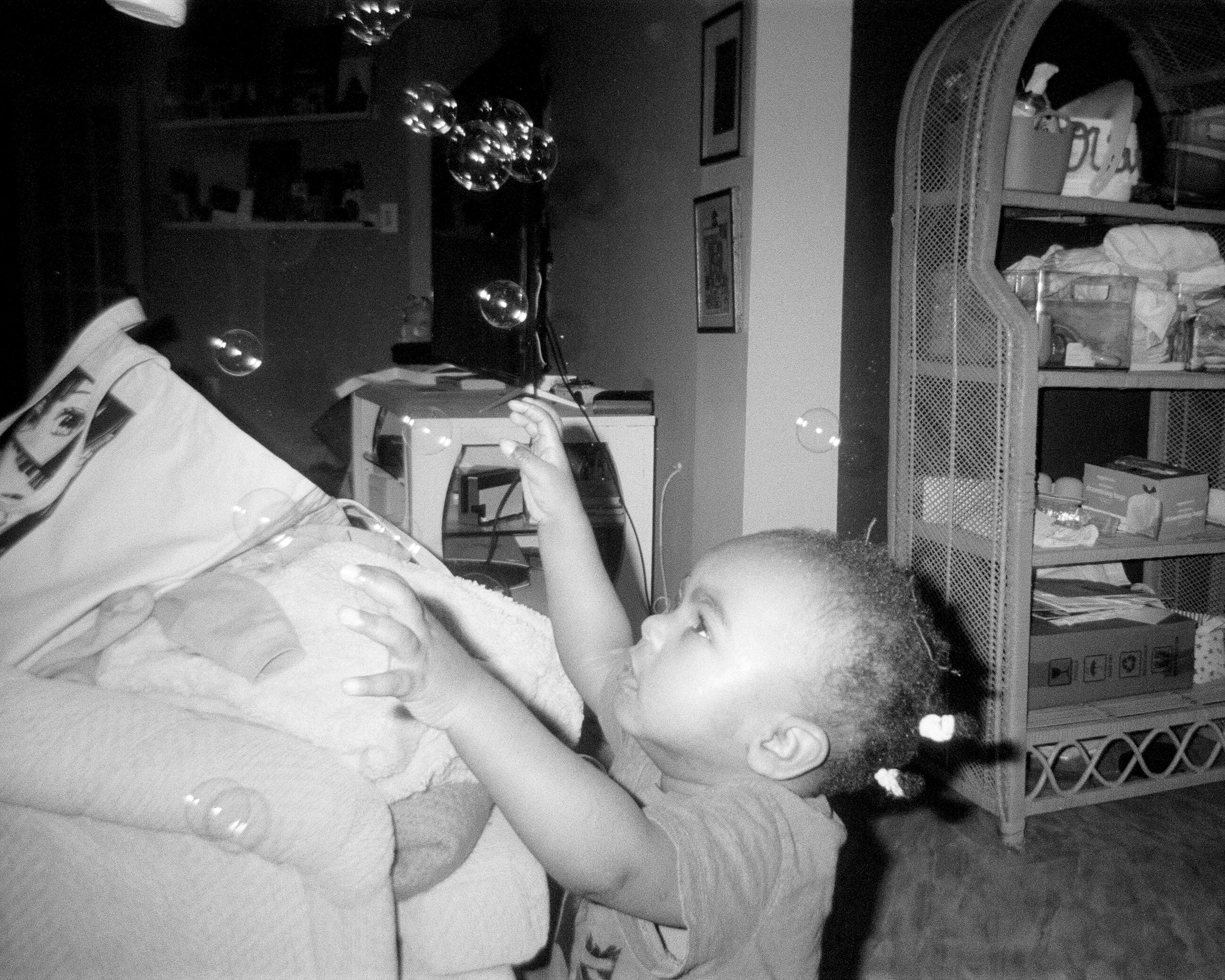 A young child reaching for bubbles indoors, with shelves and a chair in the background.