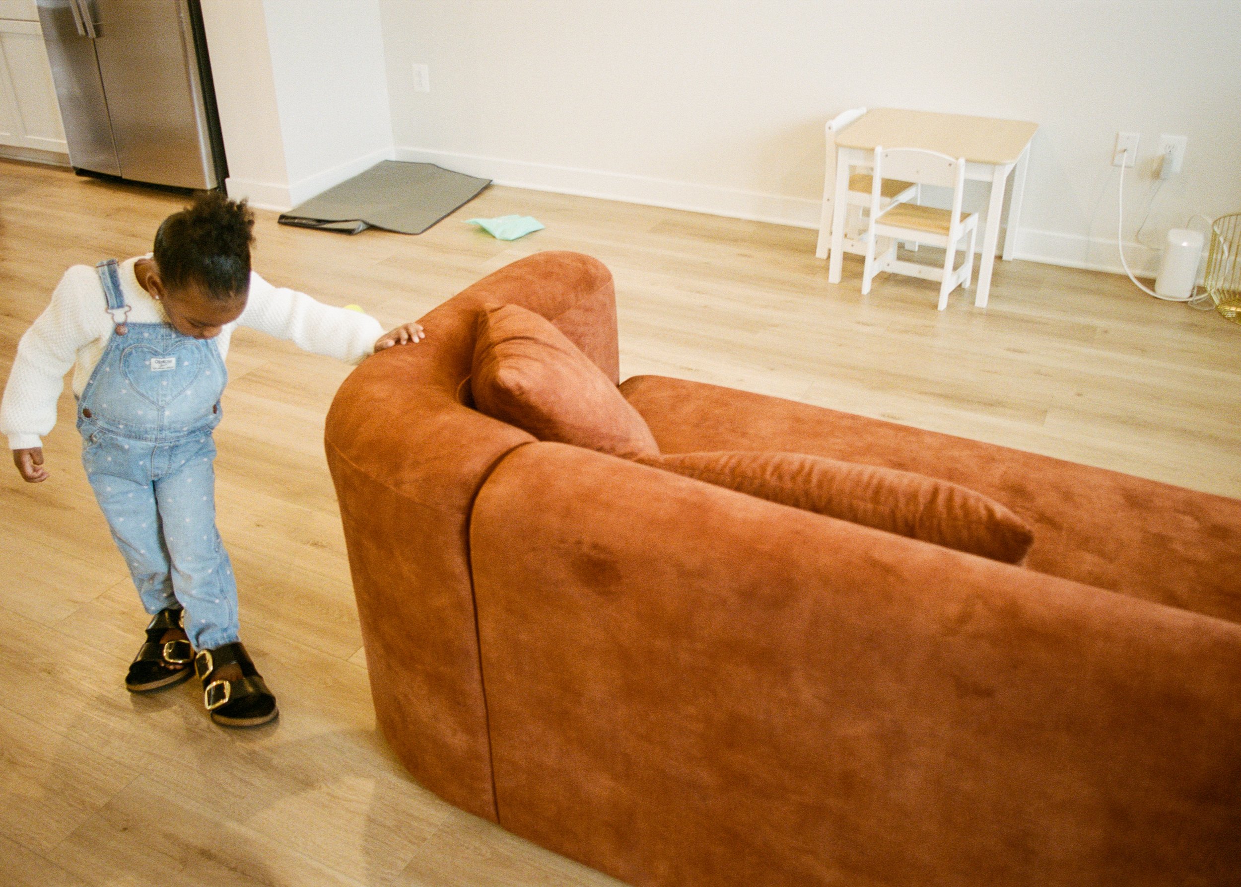 Child in denim overalls touching a brown sofa in a living room with wooden floors, small table and chairs in the background.