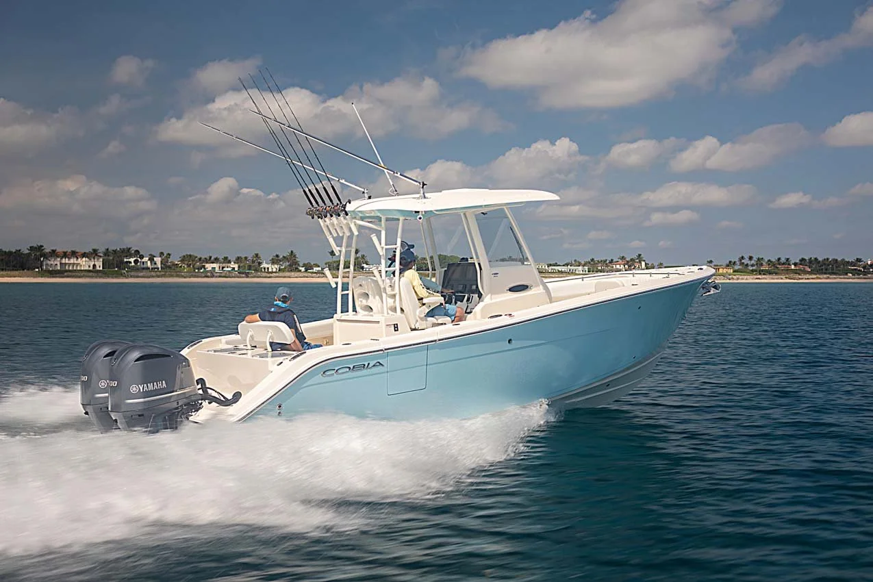 A light blue Yamaha Cobia motorboat speeding on water with three people on board, two sitting and one steering, under a partly cloudy sky with a shoreline in the background.