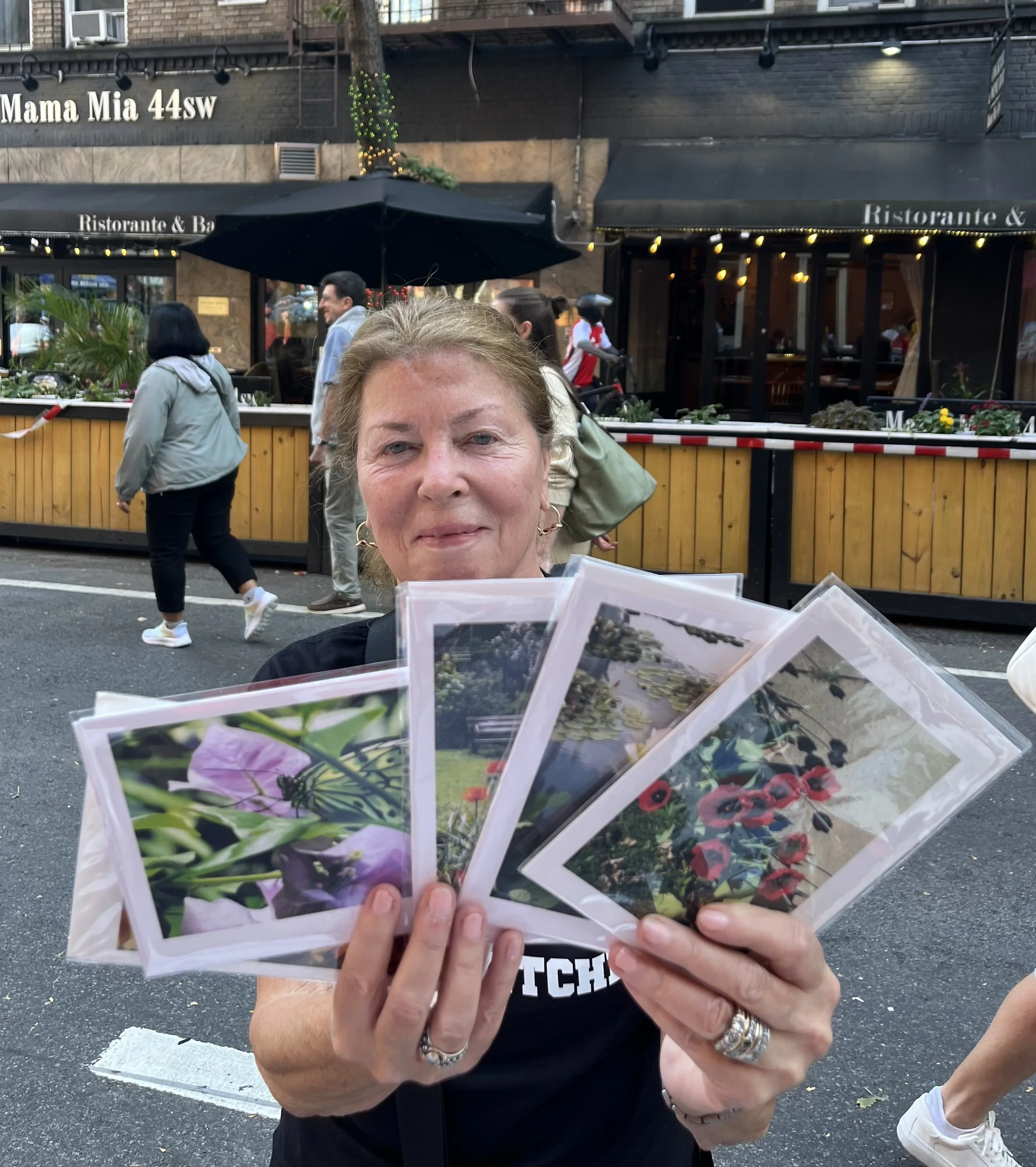 A woman holding several JanetsArthouse cards in front of her that she bought at a NYC street fair  during an outdoor event with people walking and a restaurant in the background.
