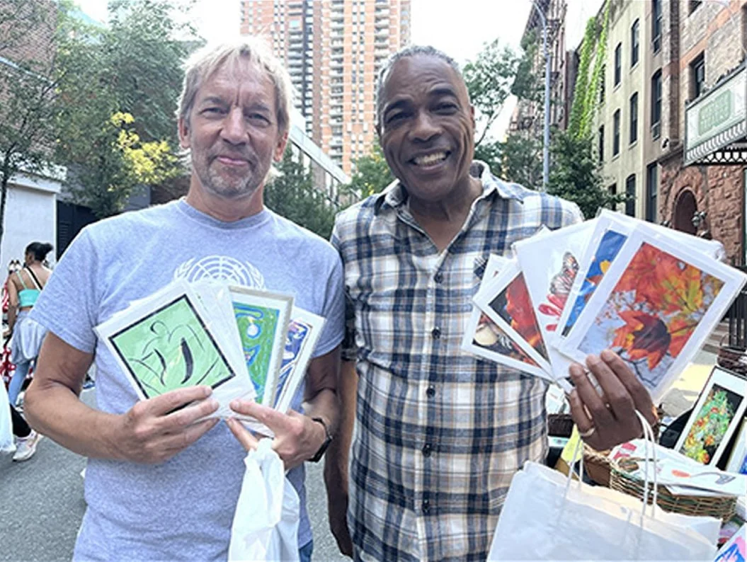 Two men standing outdoors in an urban setting, holding art prints and smiling at the camera. One man has gray hair and a beard, wearing a gray t-shirt, and the other is bald, wearing a plaid shirt. There are buildings and trees in the background.
