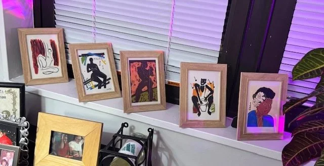 A row of five framed artworks on a windowsill, featuring various abstract and illustrative designs, with a yellow-framed photograph below and some decorative items in front.