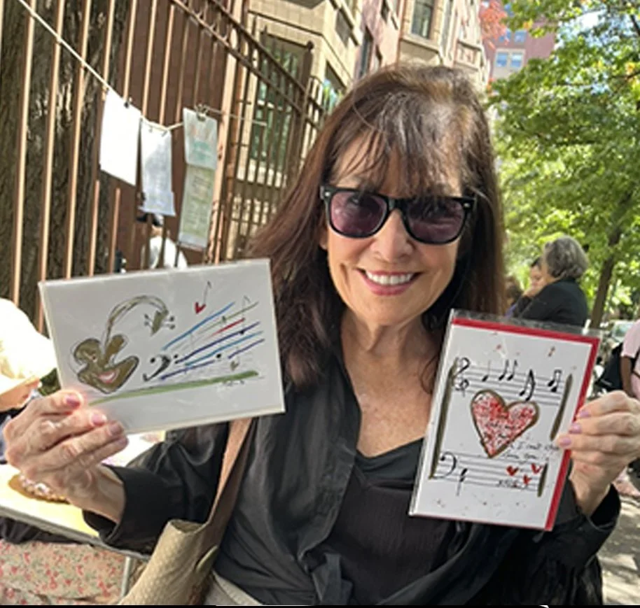 A smiling woman wearing sunglasses and a black jacket is holding two handmade cards with colorful musical and heart designs outdoors on a sunny day.